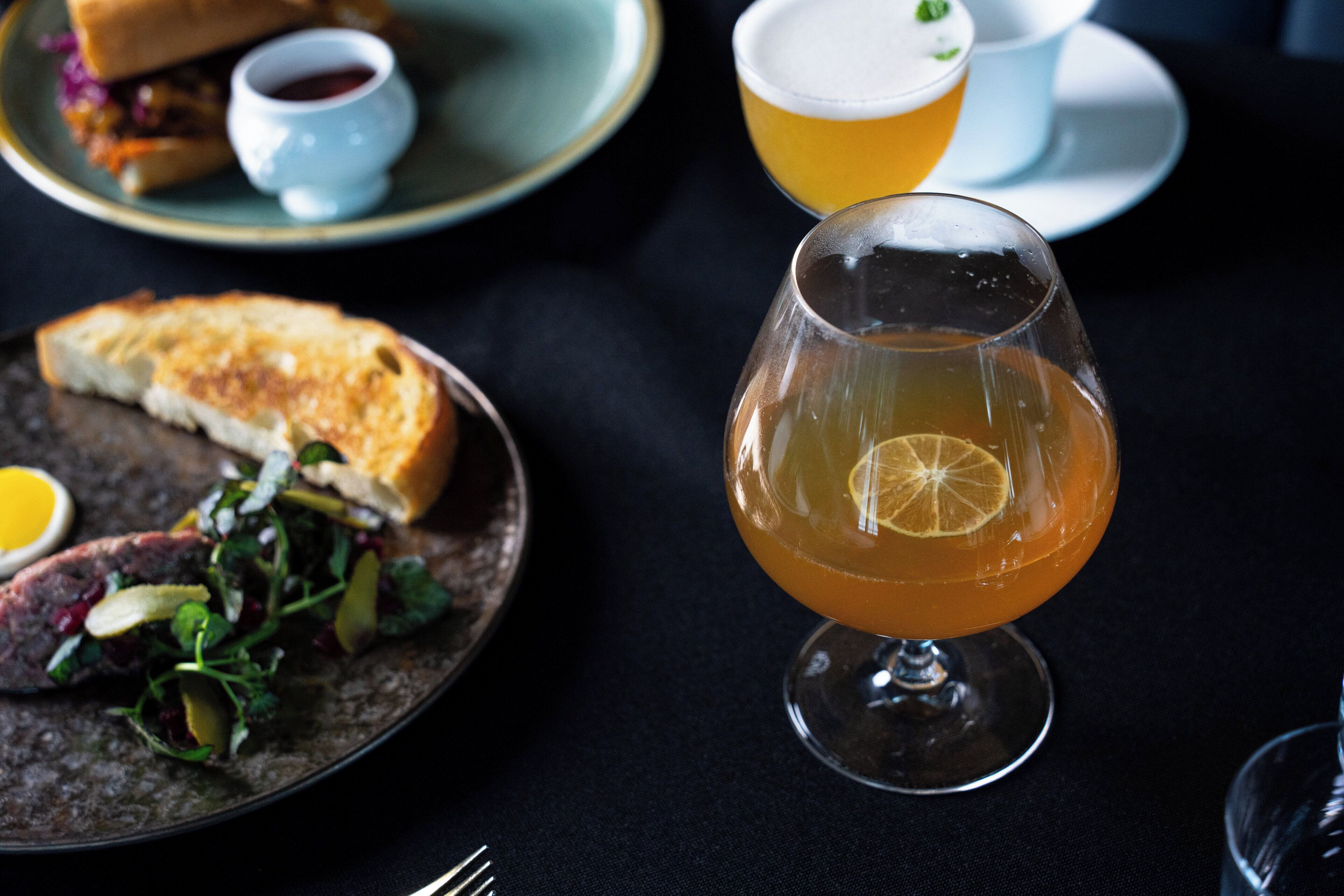 A dining table with a plate of food, a glass of amber-colored drink, and a cup of beer.