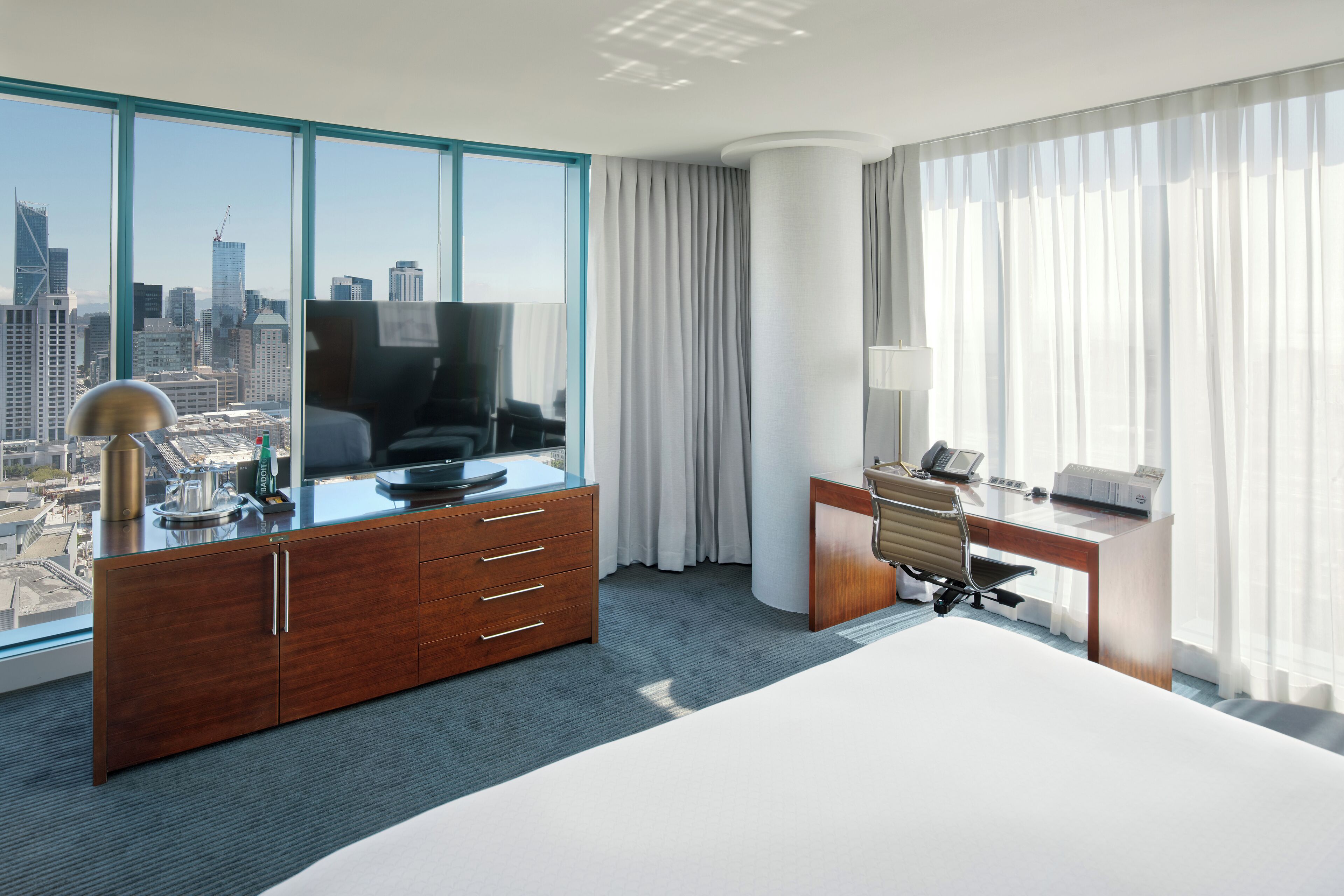 Ein modernes Hotelzimmer mit einem großen Bett, einem Schreibtisch mit Stuhl, einem Fernseher und Blick auf die Stadt durch große Fenster.