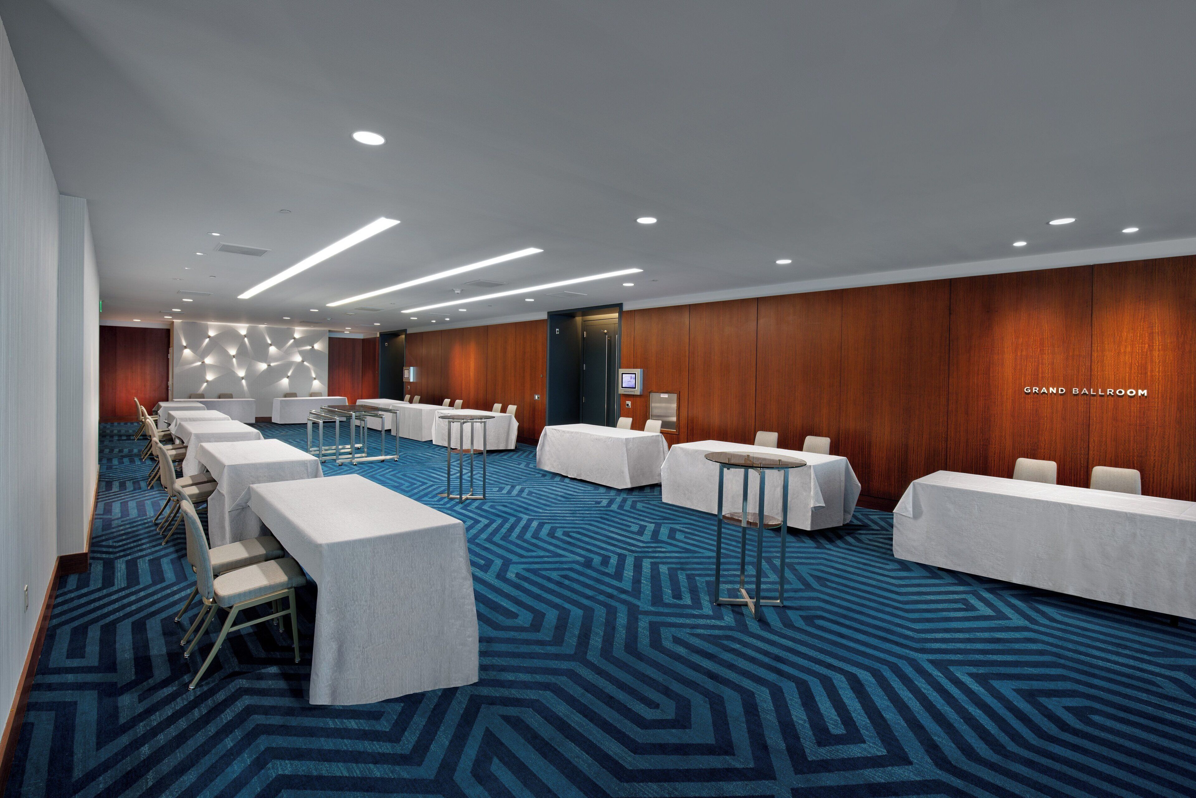 Un salón de conferencias con varias mesas y sillas, alfombra estampada y paneles de madera en las paredes.