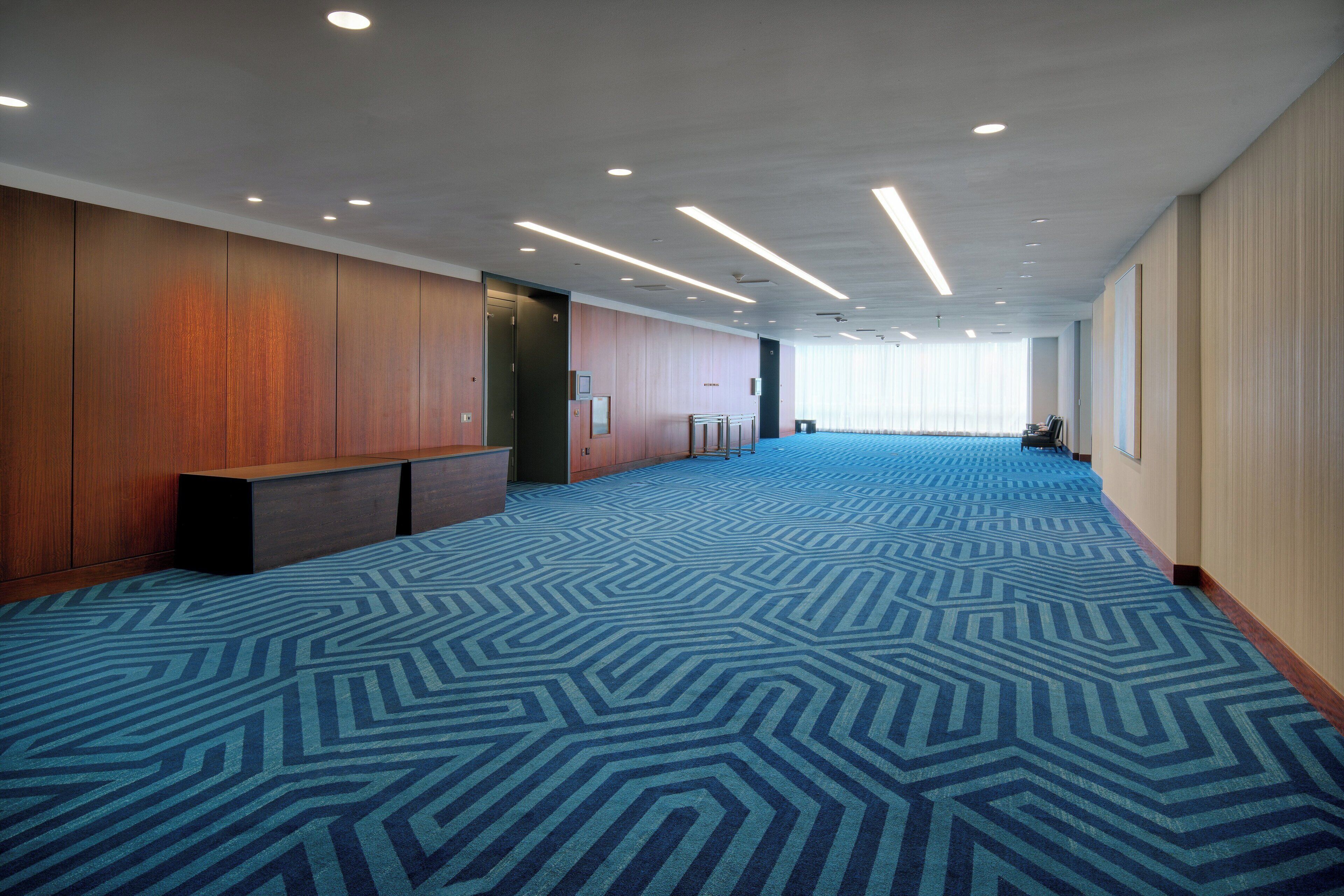 A spacious hotel lobby with a patterned carpet, wooden paneling, and a long corridor leading to a conference room.