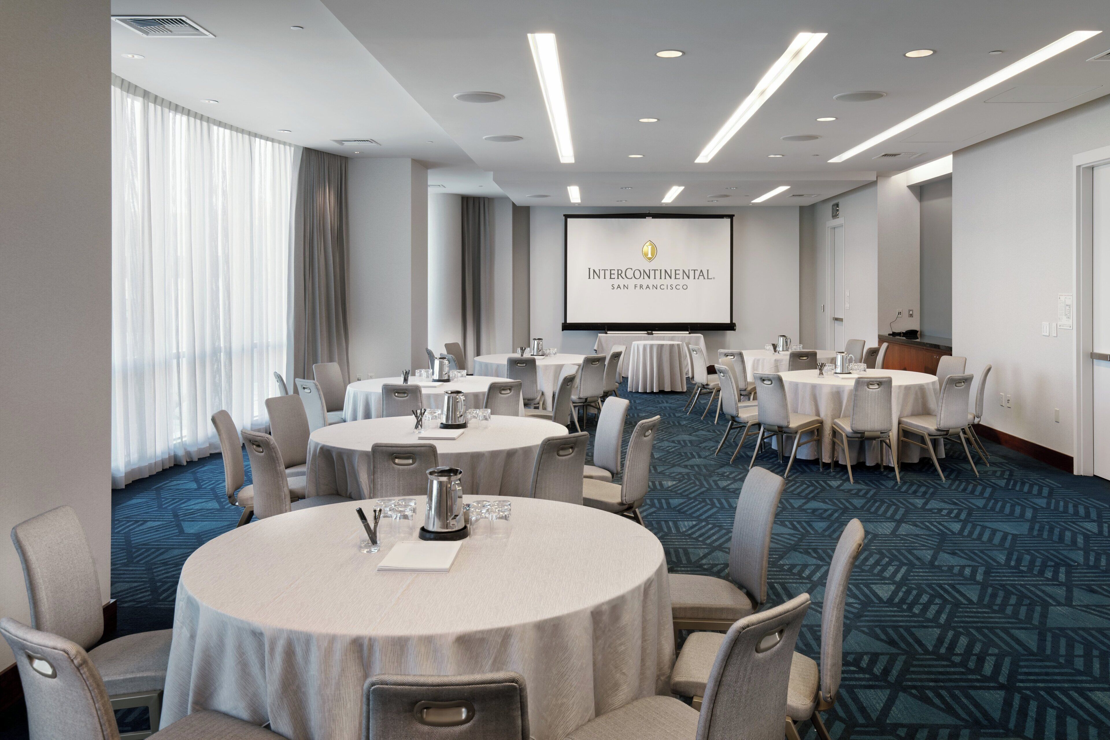 A conference room with multiple round tables, chairs, and a projection screen displaying 'Intercontinental San Francisco'.