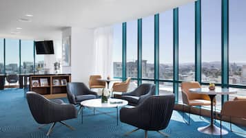 A modern hotel lobby with large windows, comfortable seating, a small table, and a bookshelf.