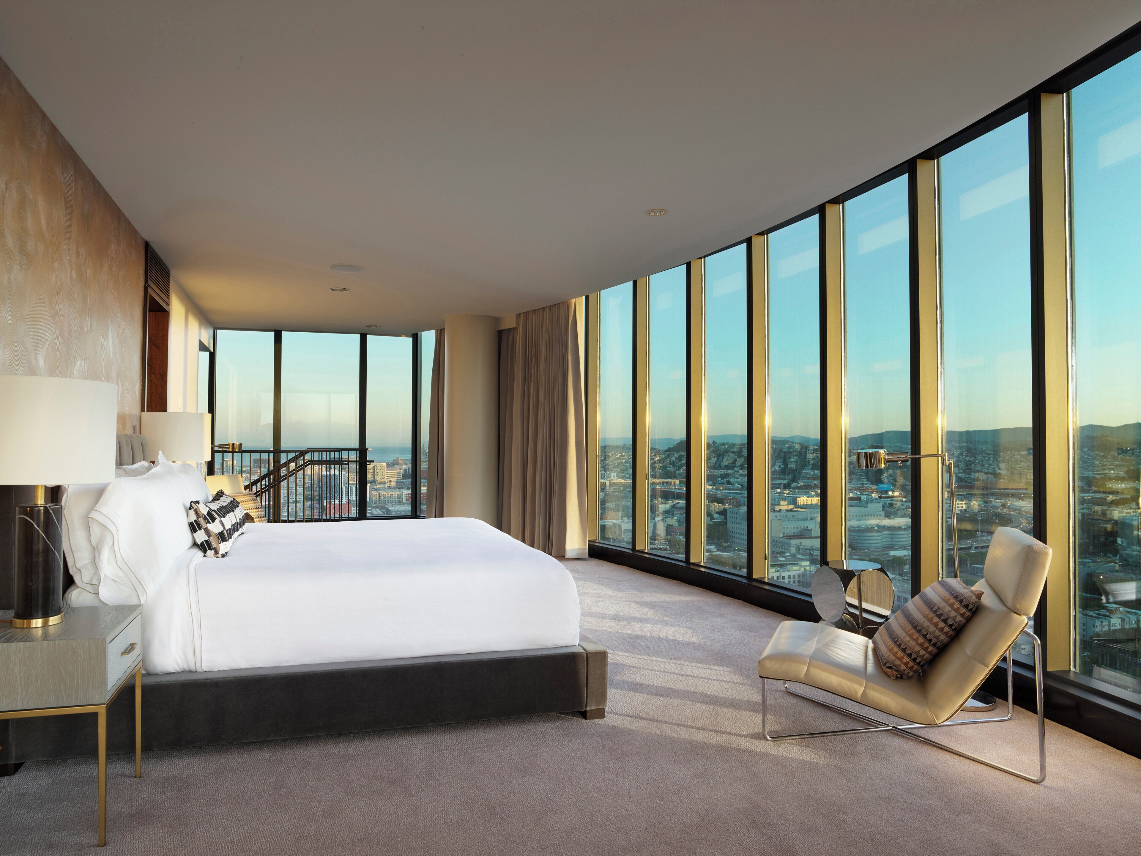 Una habitación de hotel moderna con una cama grande, una zona de estar con una silla y vistas a la ciudad a través de ventanales de techo a piso.