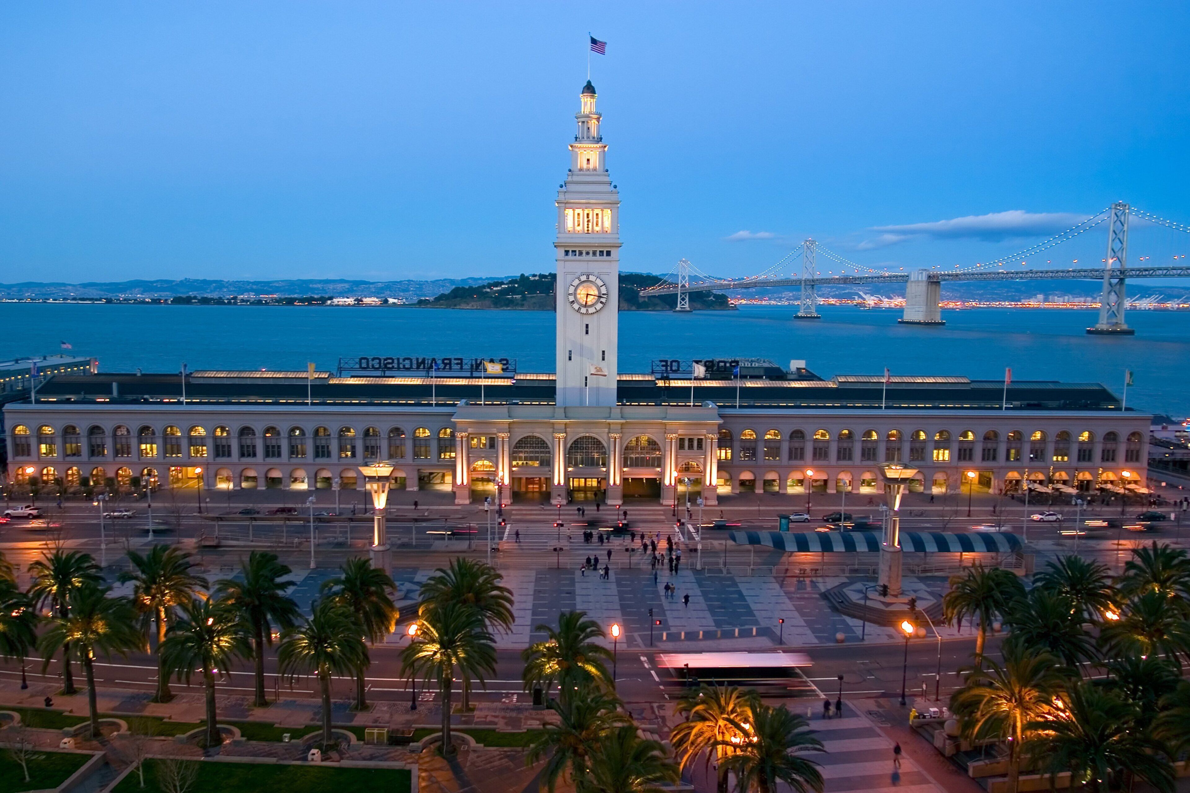 A large building with a prominent clock tower, a waterfront view, and a bridge in the background.