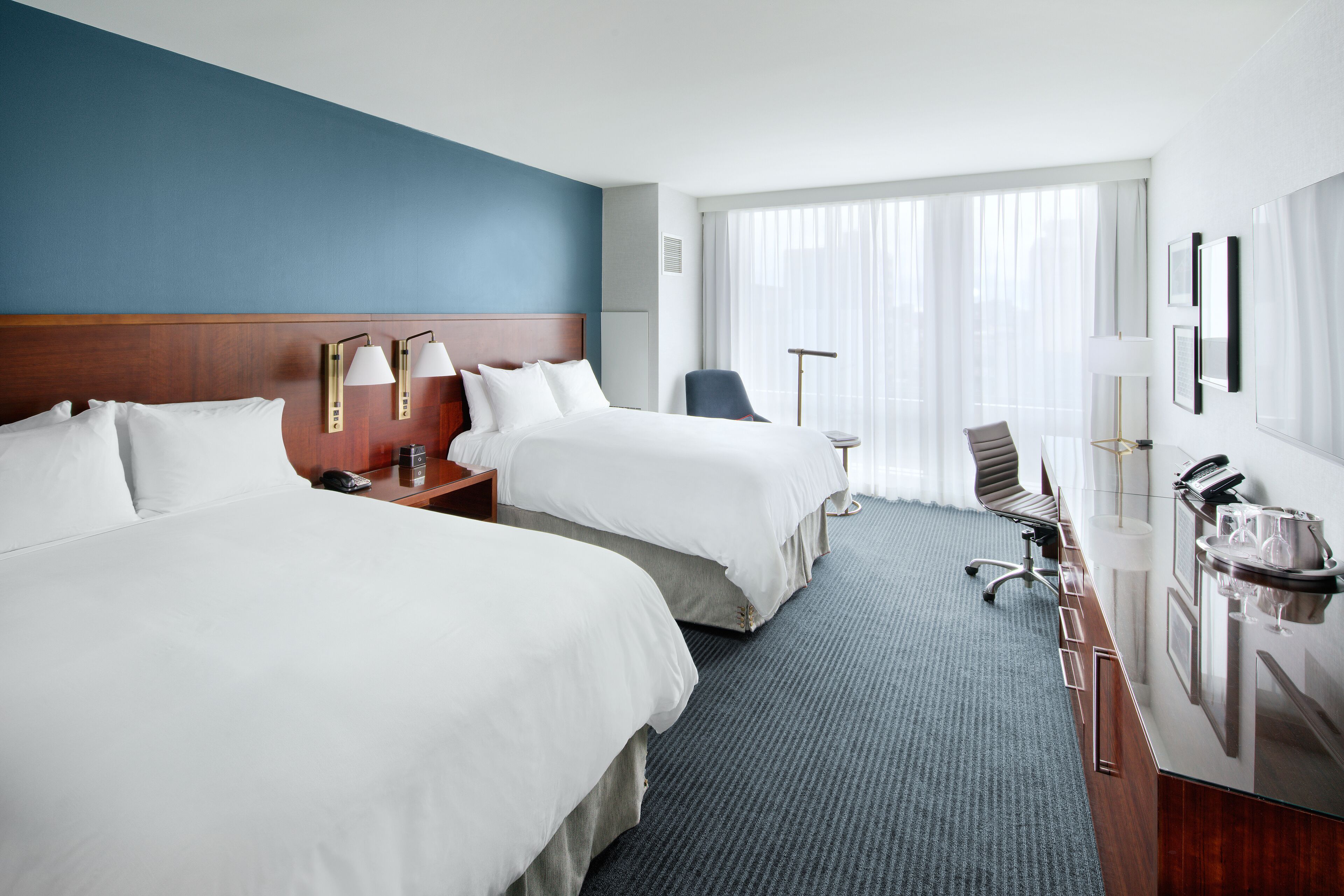 Ein Hotelzimmer mit zwei Betten, einem Schreibtisch, einem Stuhl und einem großen Fenster mit leichtem Vorhang.