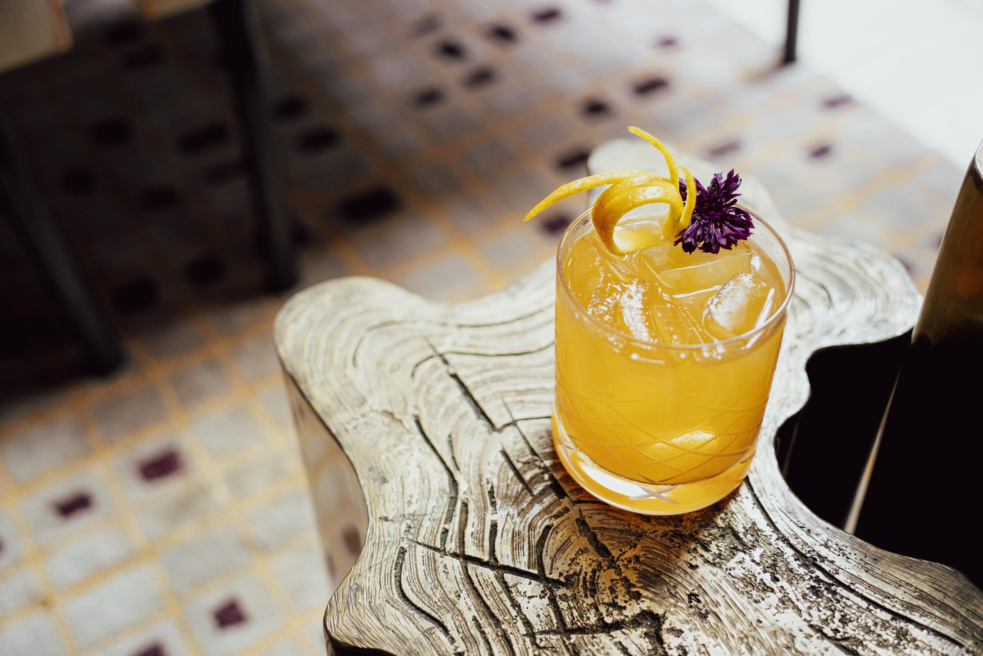 A glass of amber-colored beverage with ice cubes, garnished with a lemon peel and a purple flower, placed on a rustic wooden surface.