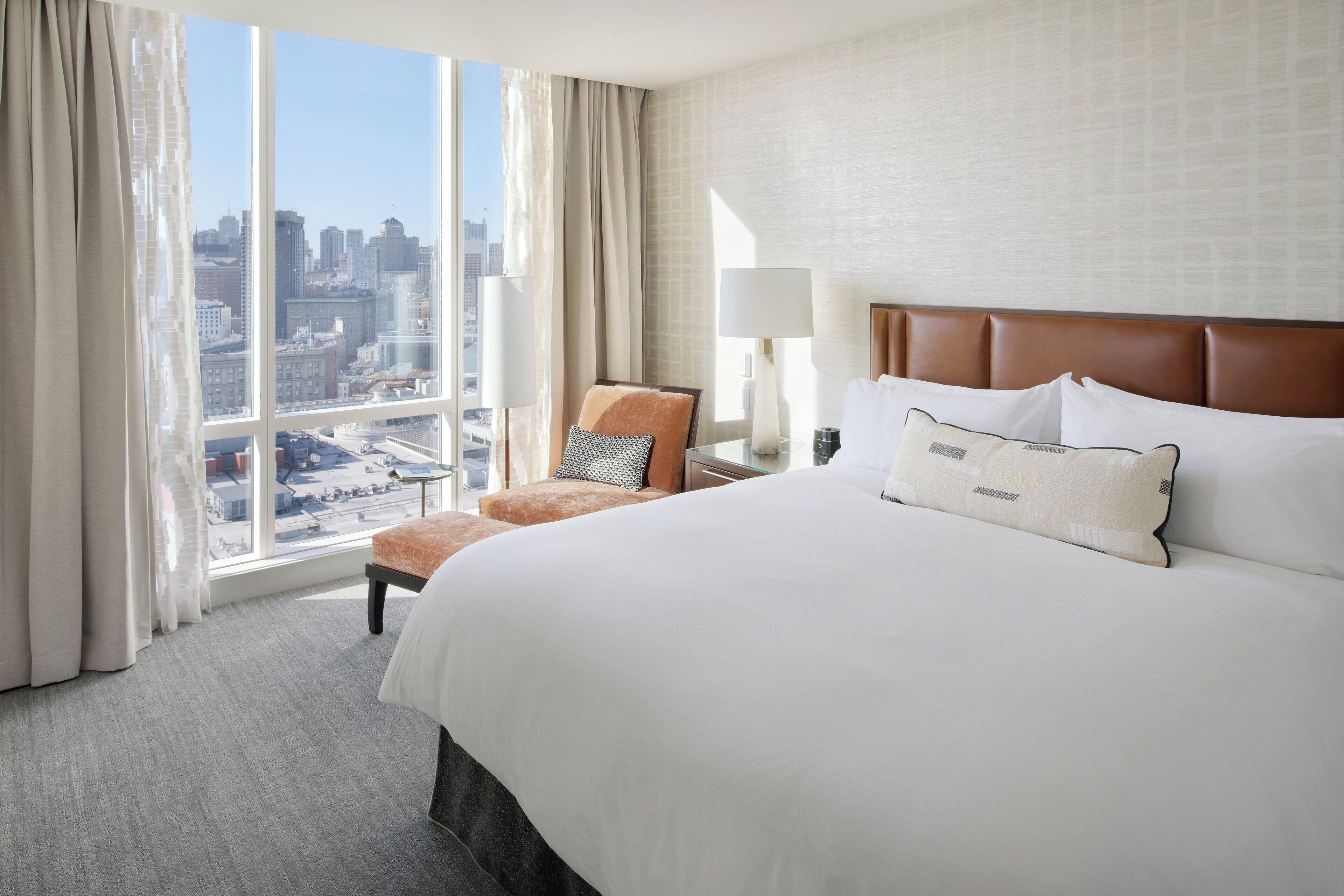 A hotel room with a large bed, a chair, a nightstand, a lamp, and a view of a cityscape through a large window.