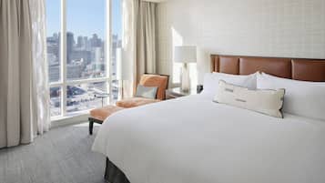 A hotel room with a large bed, a chair, a nightstand, a lamp, and a view of a cityscape through a large window.