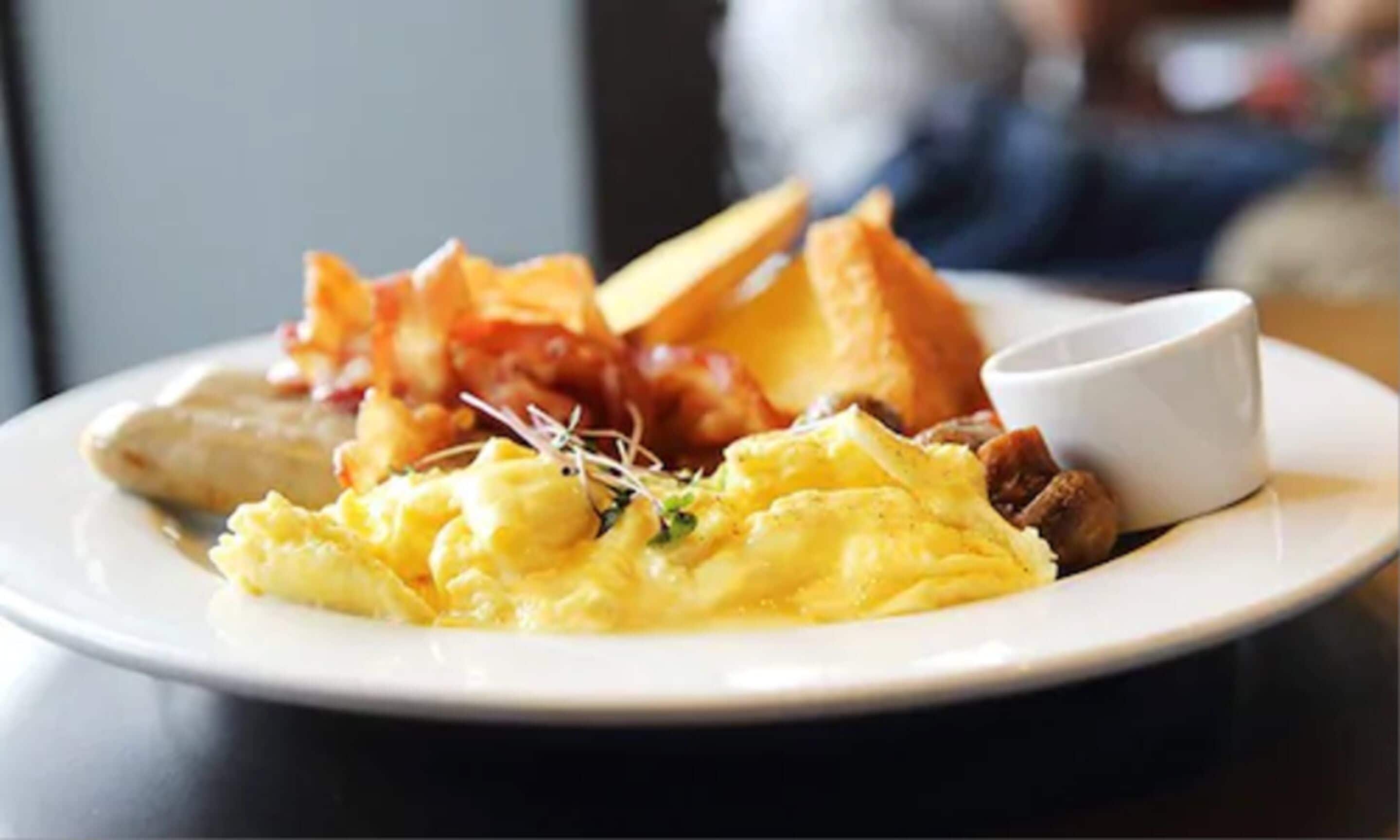 A plate of scrambled eggs, bacon, toast, and a small cup of sauce.