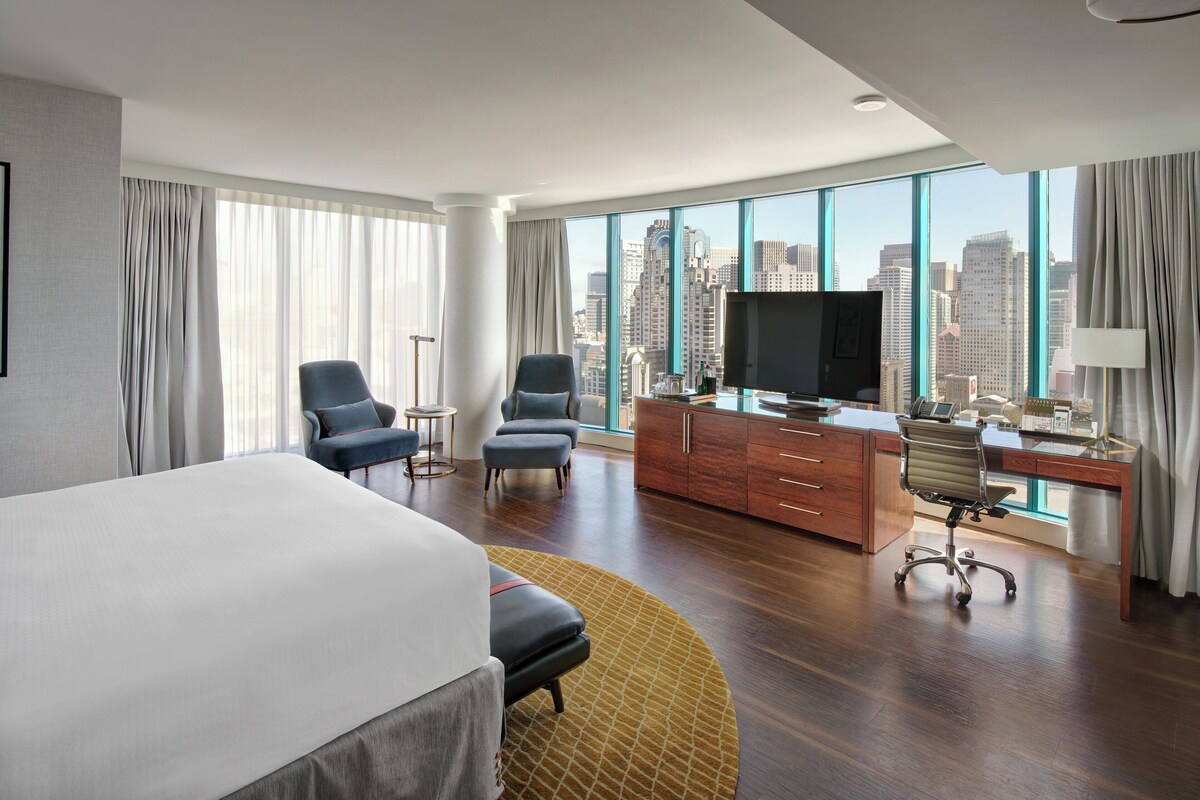 Une chambre d’hôtel avec un grand lit, deux fauteuils, un bureau avec un ordinateur, une télévision et une vue sur la ville.