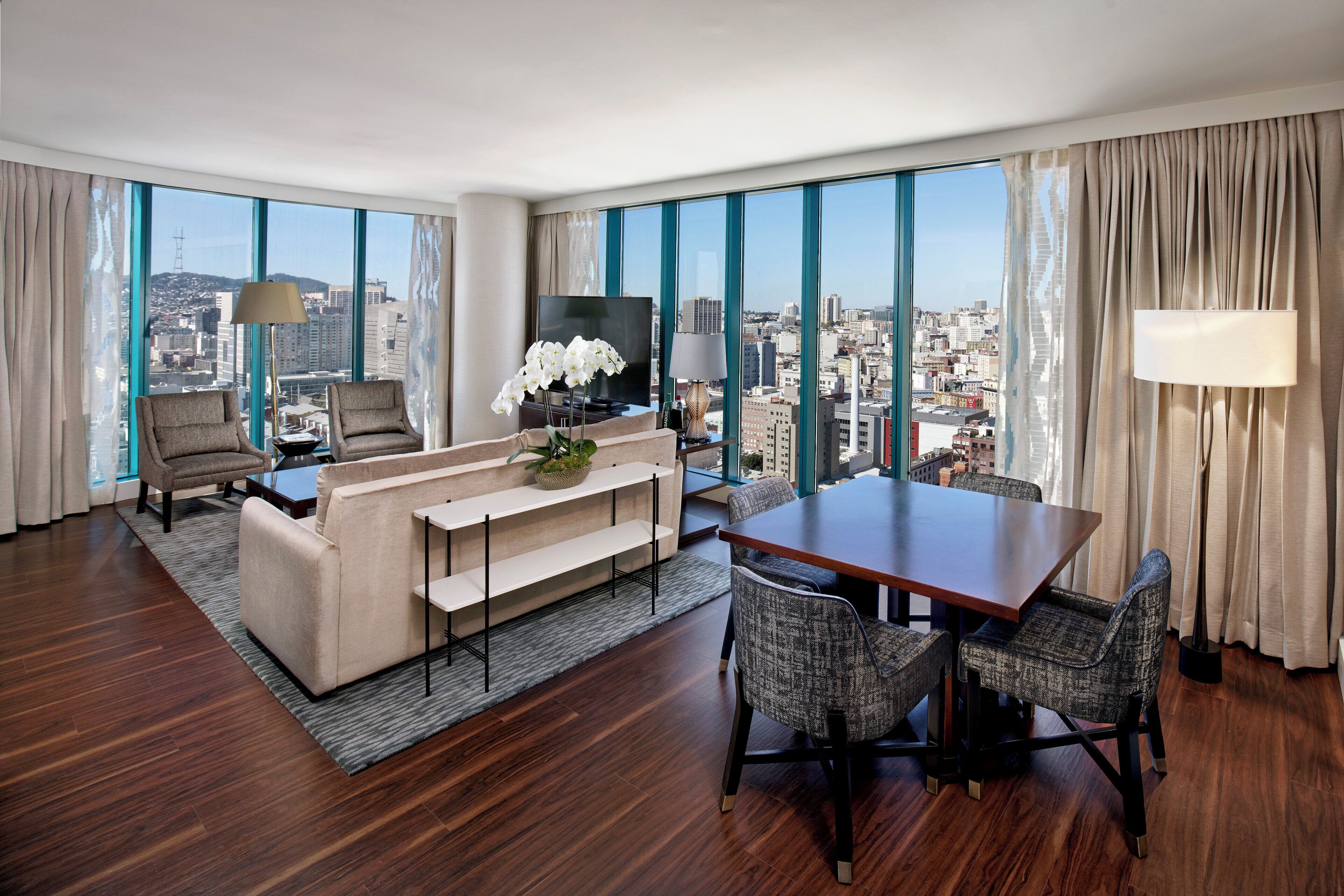 A modern hotel room with a large window, a sofa, a dining table, and a TV.