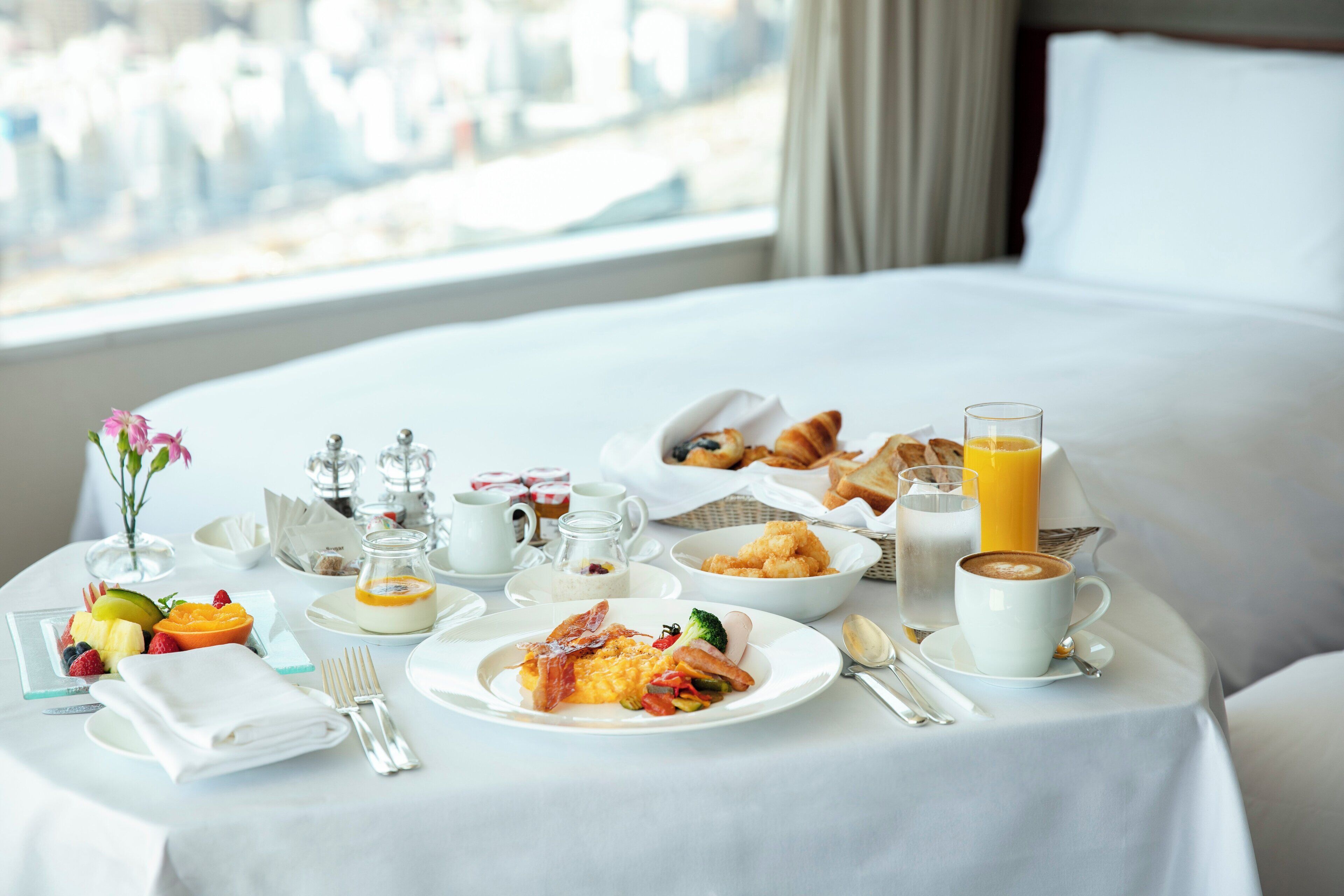 A breakfast setup on a bed with a city view, including plates of food, a glass of orange juice, and a cup of coffee.