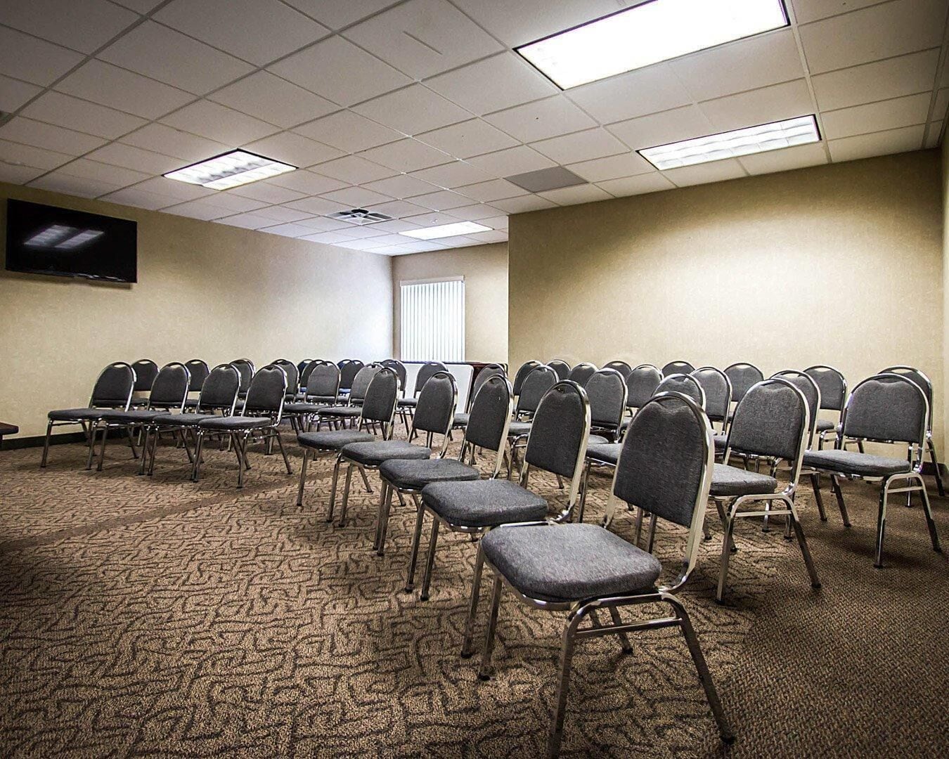 A conference room with rows of chairs, carpeted floor, ceiling lights, and a TV on the wall.