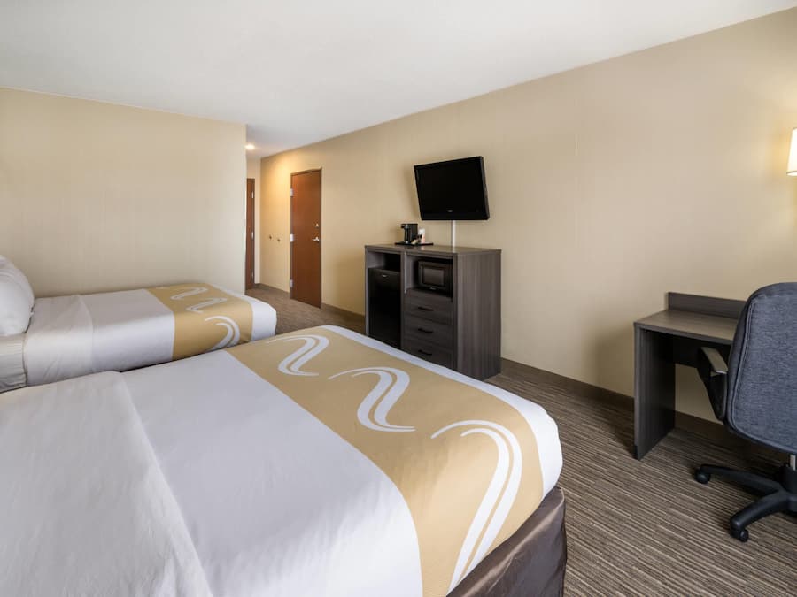 A hotel room with two beds, a desk, a chair, a television, and a lamp.