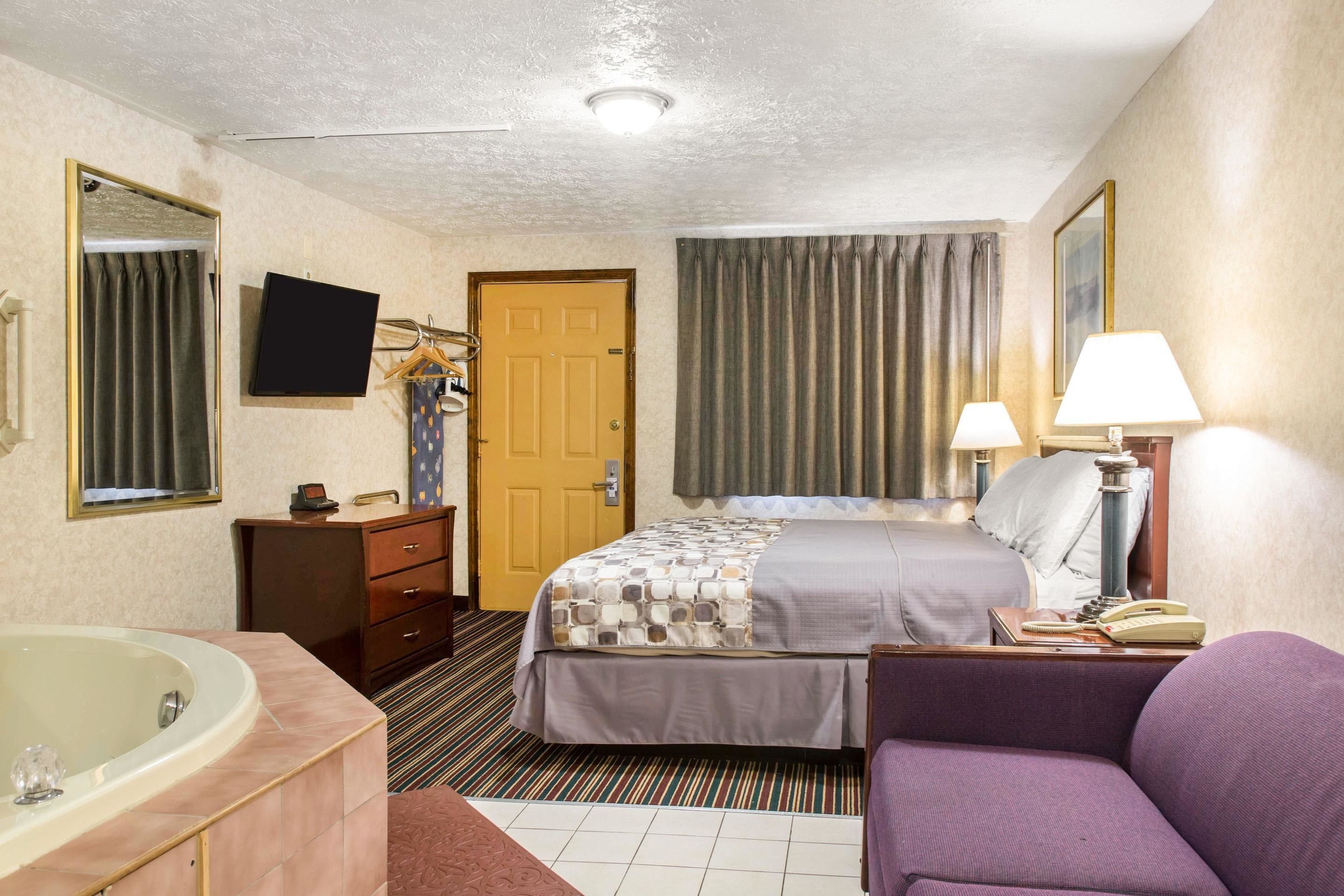 A hotel room with a bed, a desk with a lamp, a chair, a bathroom with a bathtub, and a television on the wall.