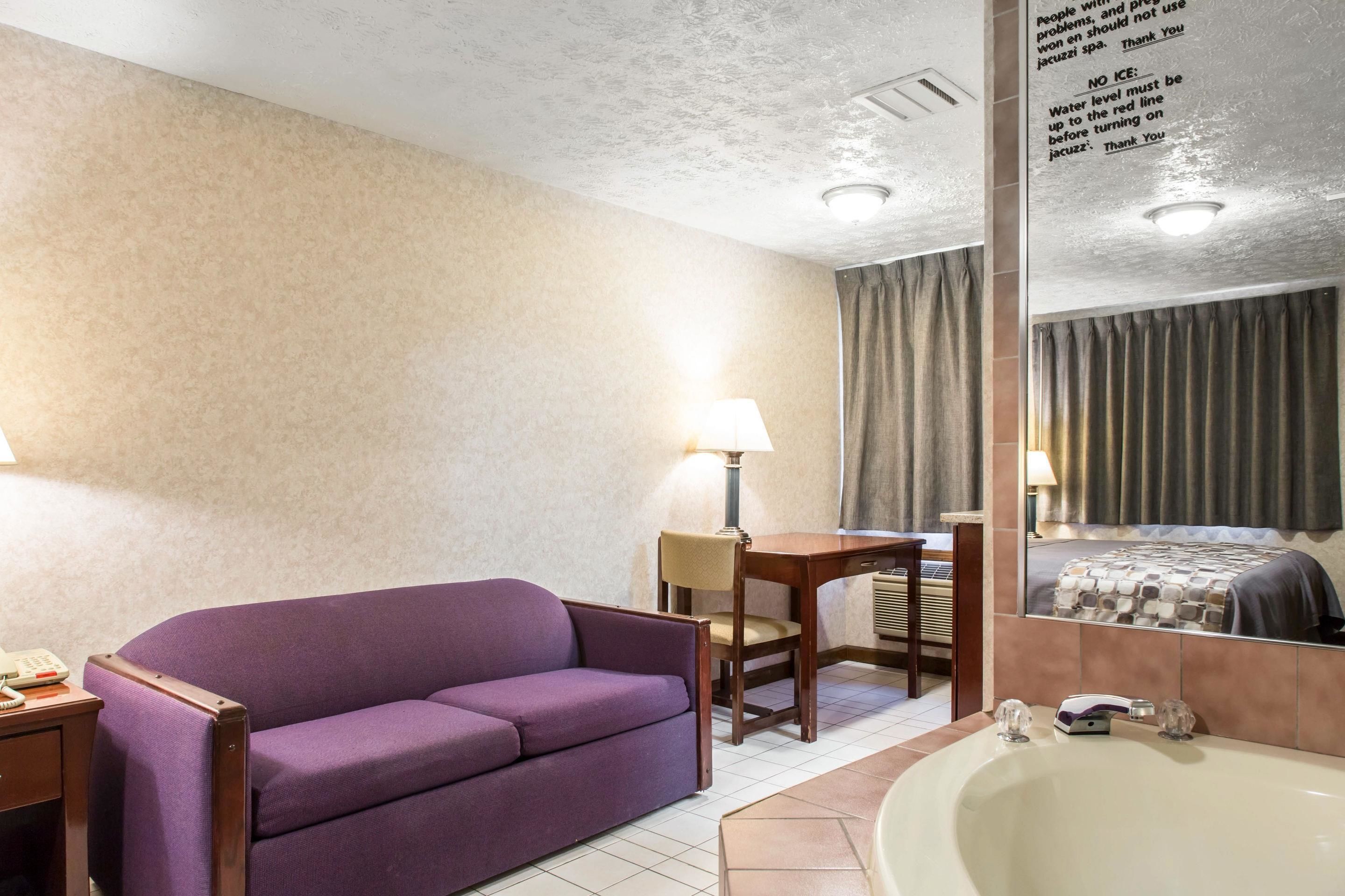 Una camera d'albergo con un divano viola, un tavolino con una lampada, uno specchio grande e una vasca da bagno.