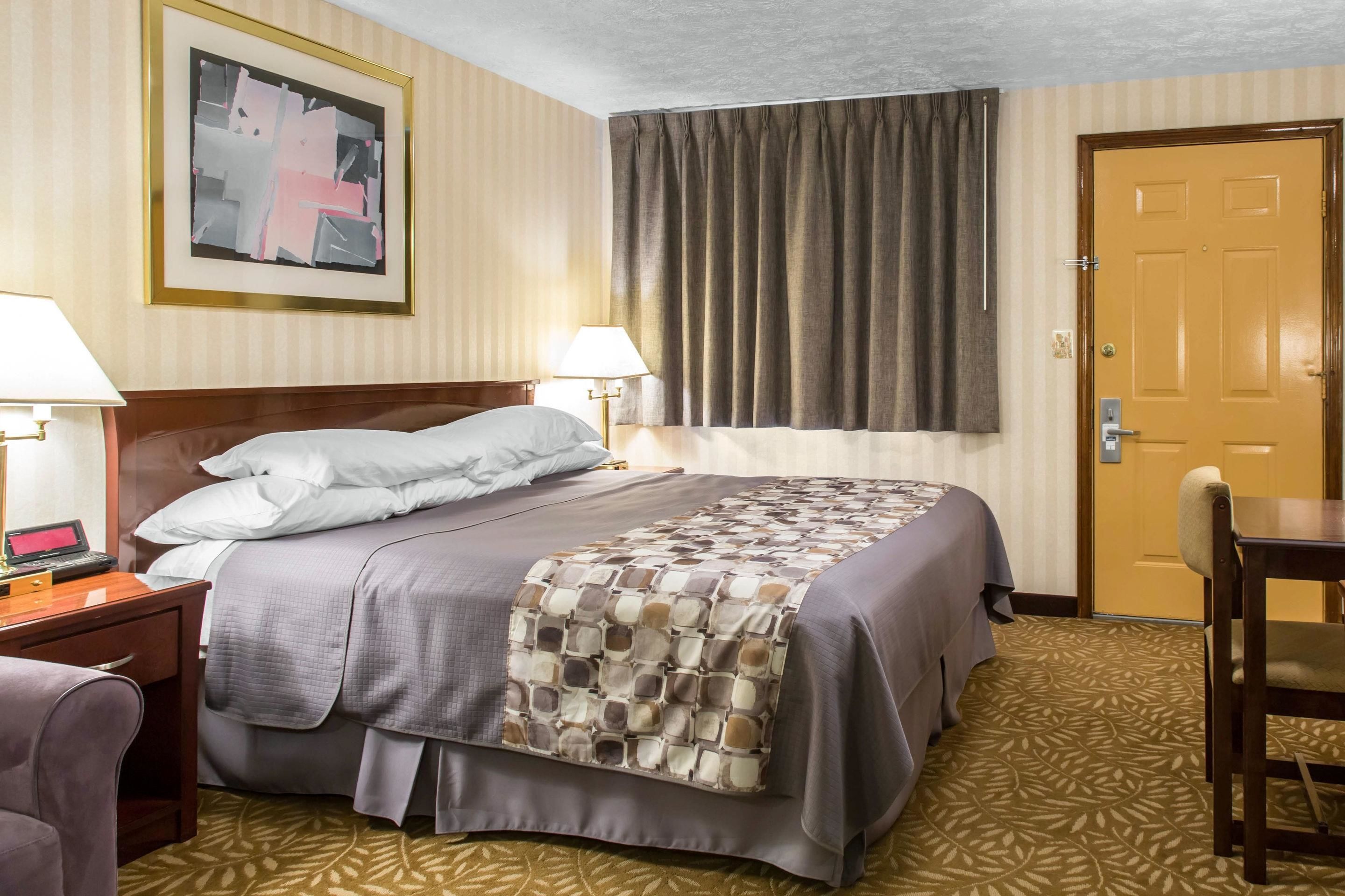 Ein Hotelzimmer mit Bett, Nachttischlampen, einem Schreibtisch mit Computer, einem Stuhl, einer Tür und einem Fenster mit Vorhängen.