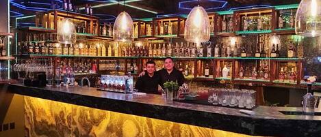 Un bar con mostrador de mármol, varias estanterías llenas de botellas de licor y lámparas colgantes.