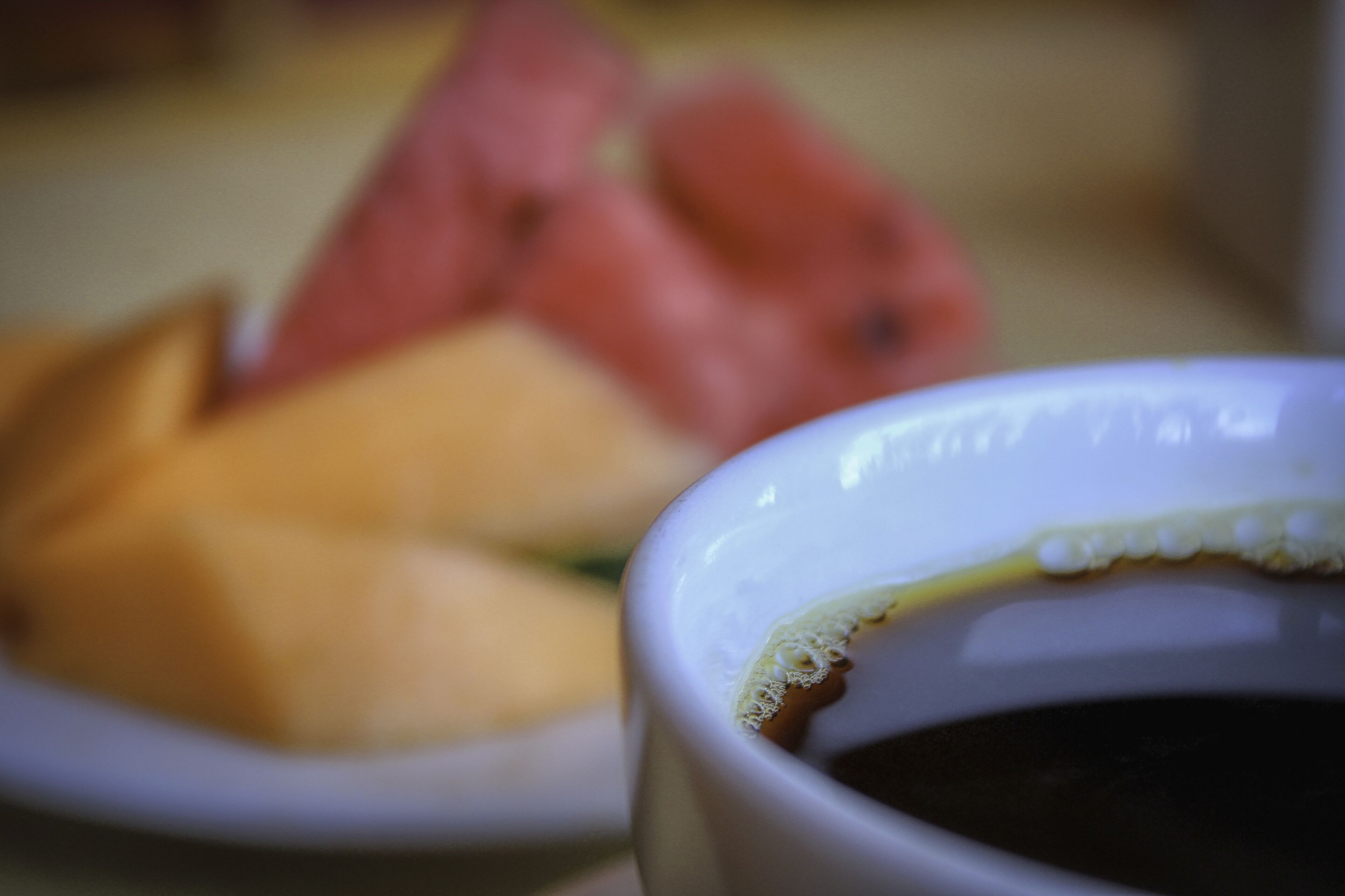 A close-up of a white ceramic cup with a beverage, possibly coffee, and a blurred background featuring a plate with food items.