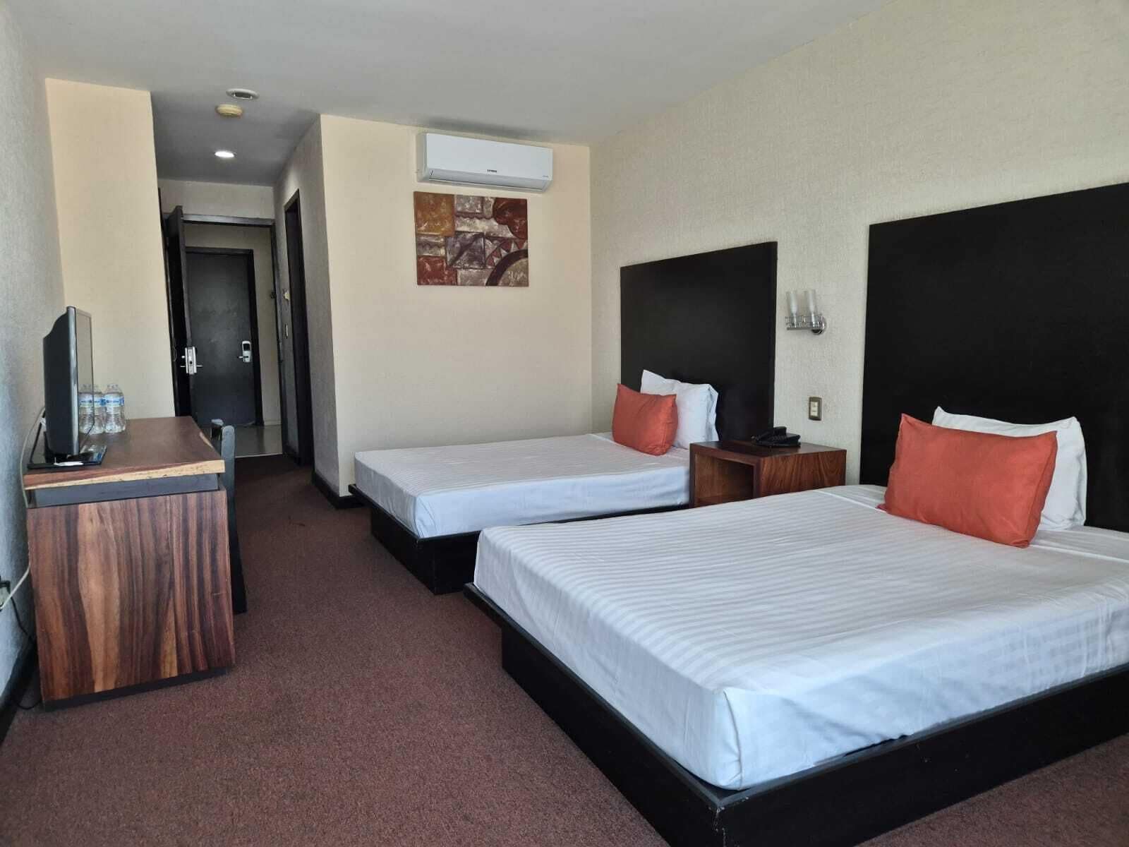 A hotel room with two beds, a desk, a TV, and an air conditioning unit.