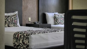 Habitación de hotel con dos camas, cada una con ropa de cama y almohadas estampadas. Hay una mesita de noche con una lámpara y una pequeña mesa entre las camas.