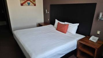 A hotel room with a large bed, a wooden nightstand with a phone, a wall-mounted light, and a painting above the bed.