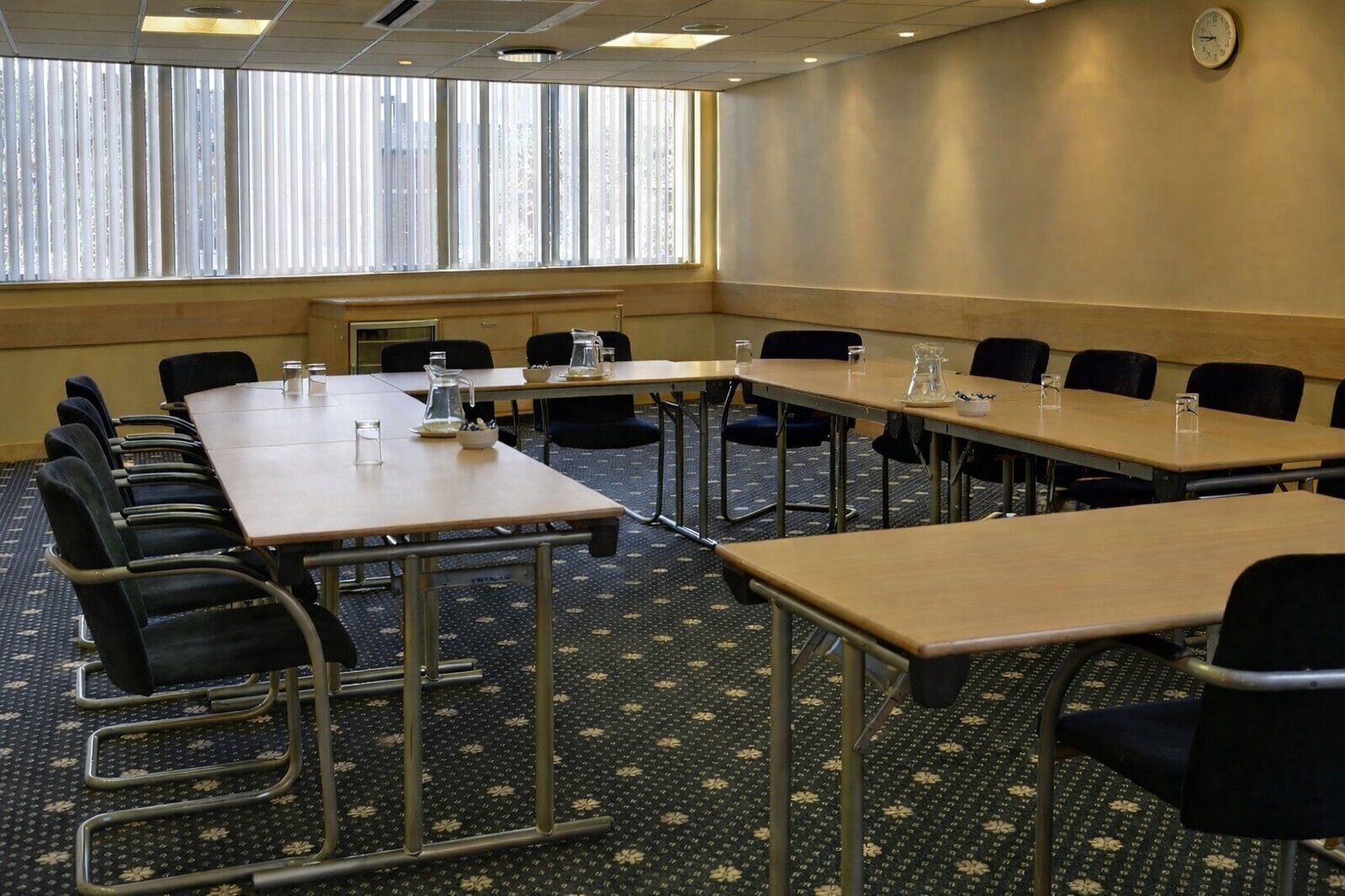 A conference room with long tables and chairs, a clock on the wall, and a window with blinds.