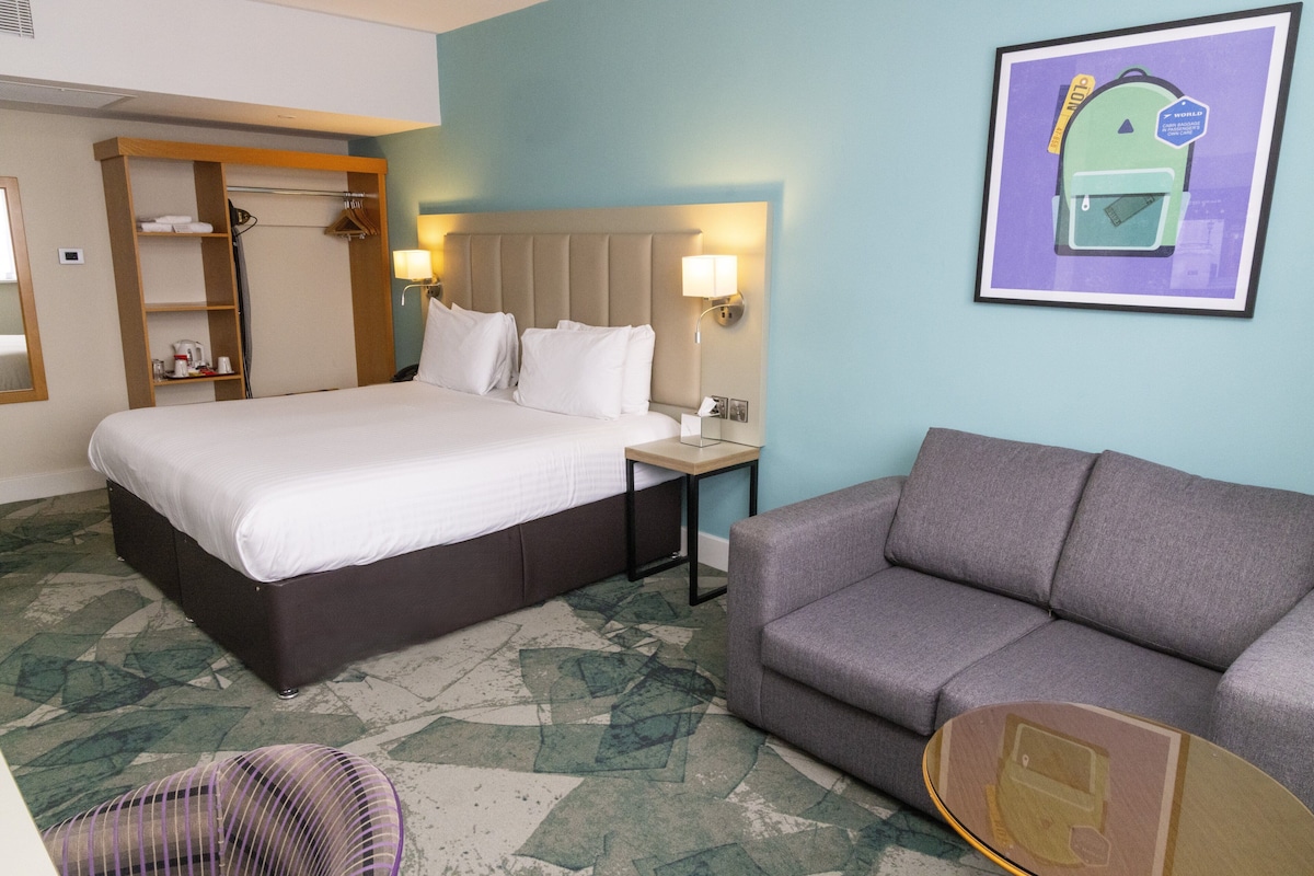 A hotel room with a bed, a gray sofa, a small table, a chair, and a framed picture on the wall.