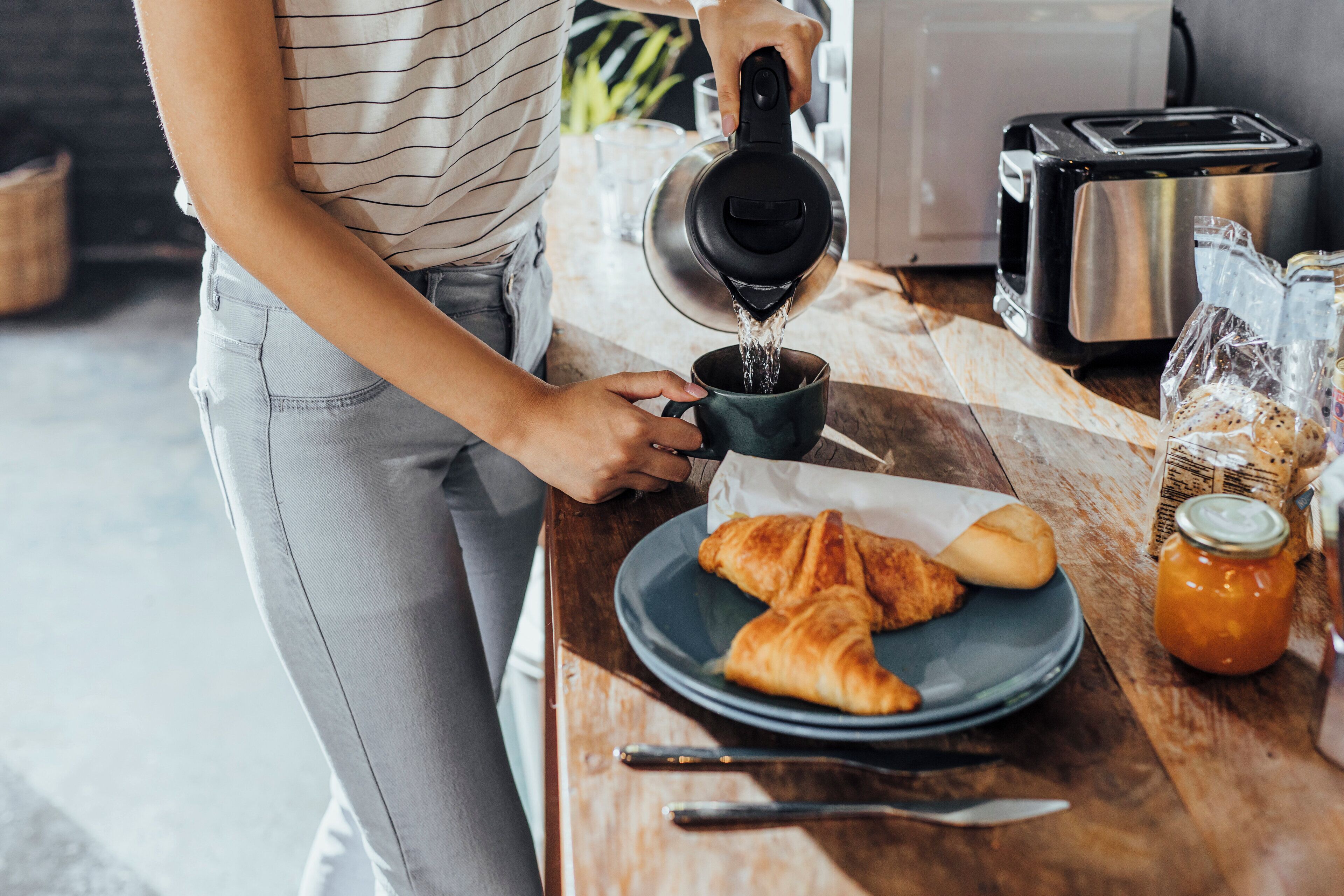 Una persona sirviendo agua en una taza sobre una mesa de madera con una tostadora, croissants y un frasco de mermelada.