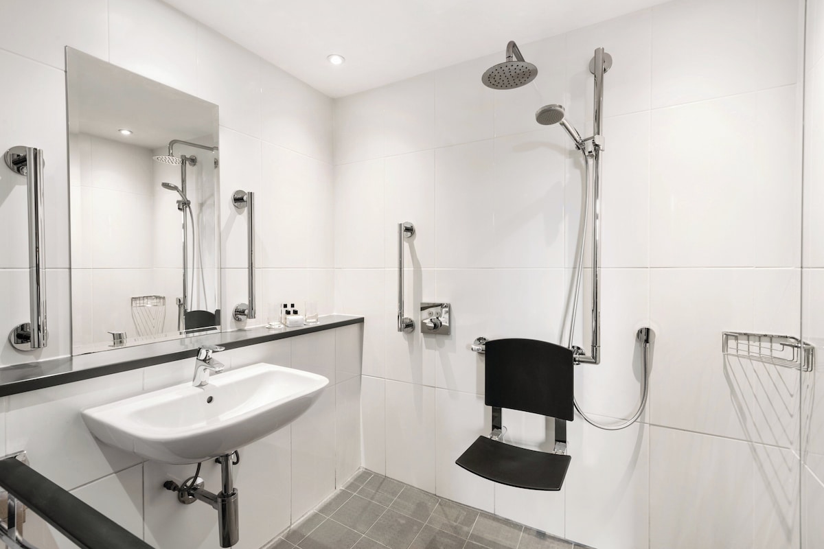 A modern bathroom with a white sink, a mirror, a shower with a handheld showerhead, and a black chair.