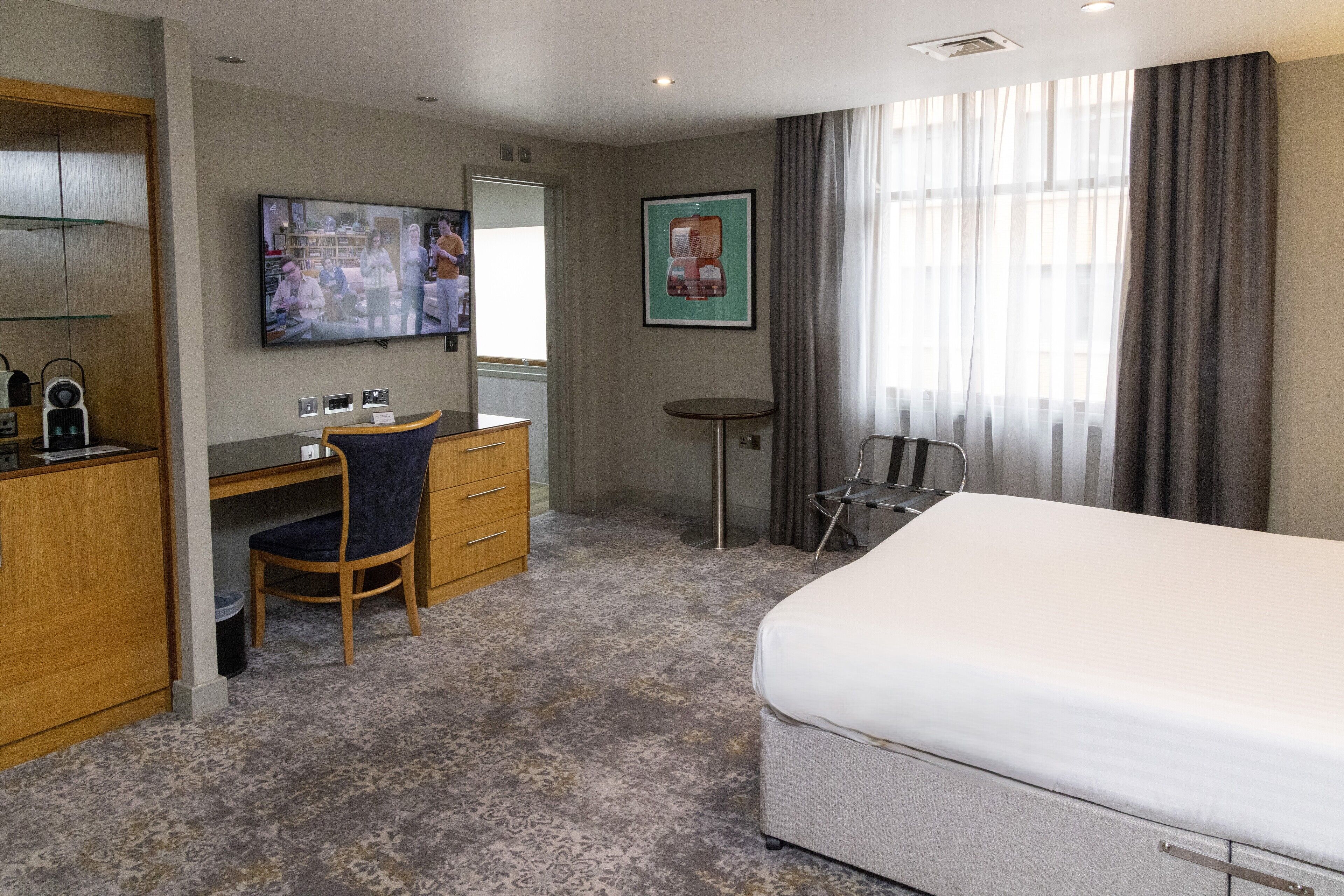 Habitación de hotel con cama, escritorio, silla, televisión y ventana con cortinas.
