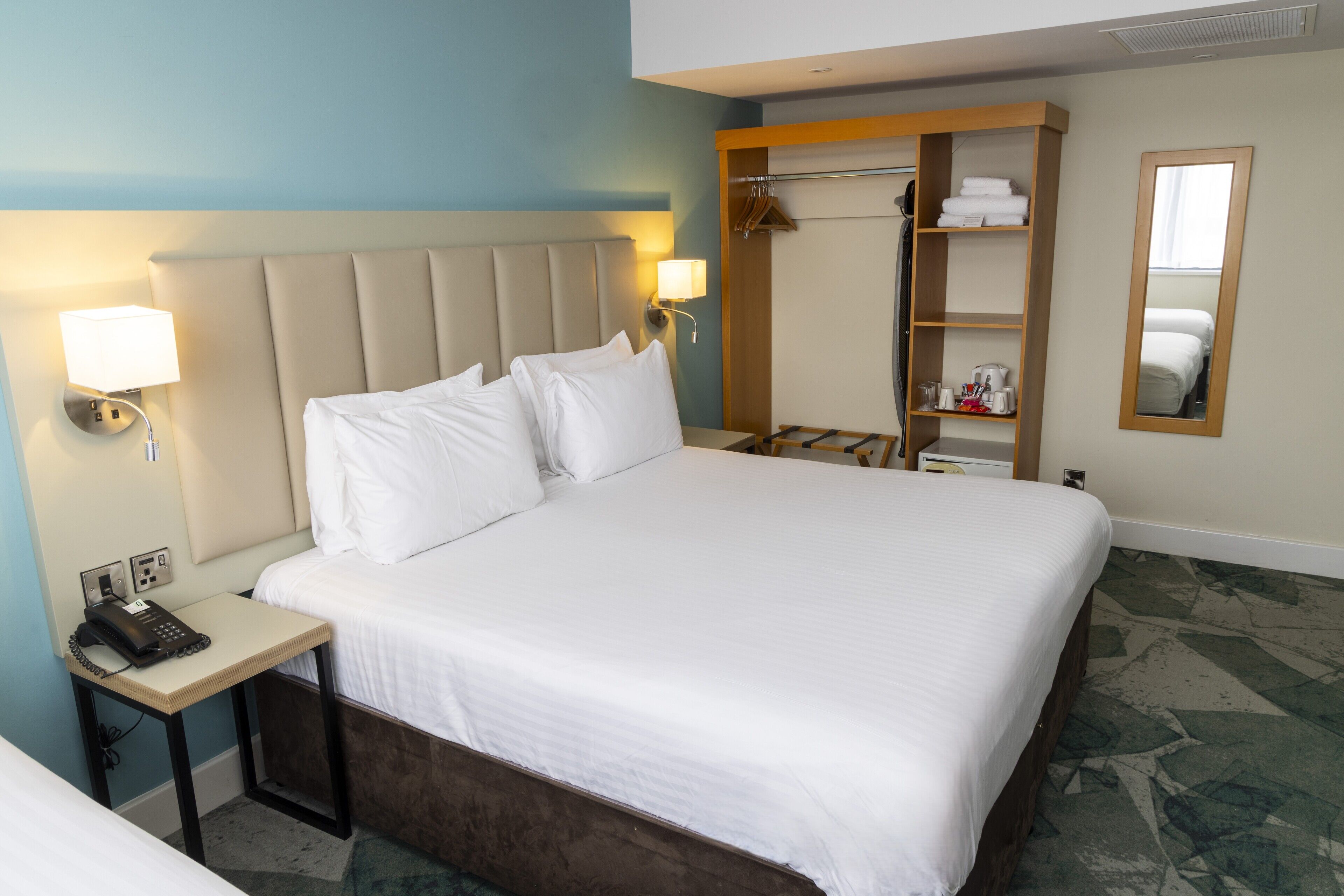 A hotel room with a large bed, bedside table with a phone, a wardrobe, and a mirror.