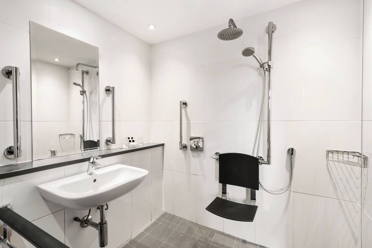 Une salle de bain moderne avec un lavabo blanc, un miroir, une douche équipée d’un pommeau de douche à main et une chaise noire.