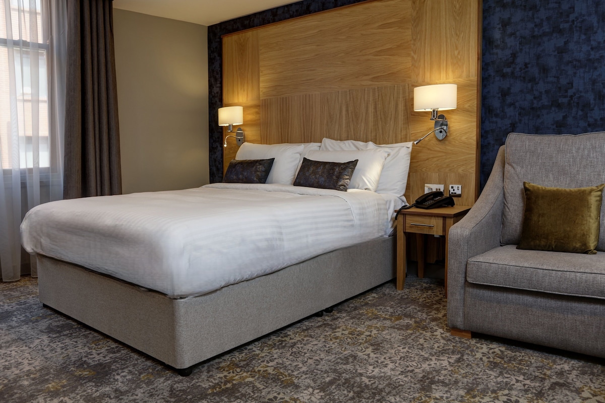 A hotel room with a large bed, two bedside lamps, a desk with a telephone, and a grey armchair.