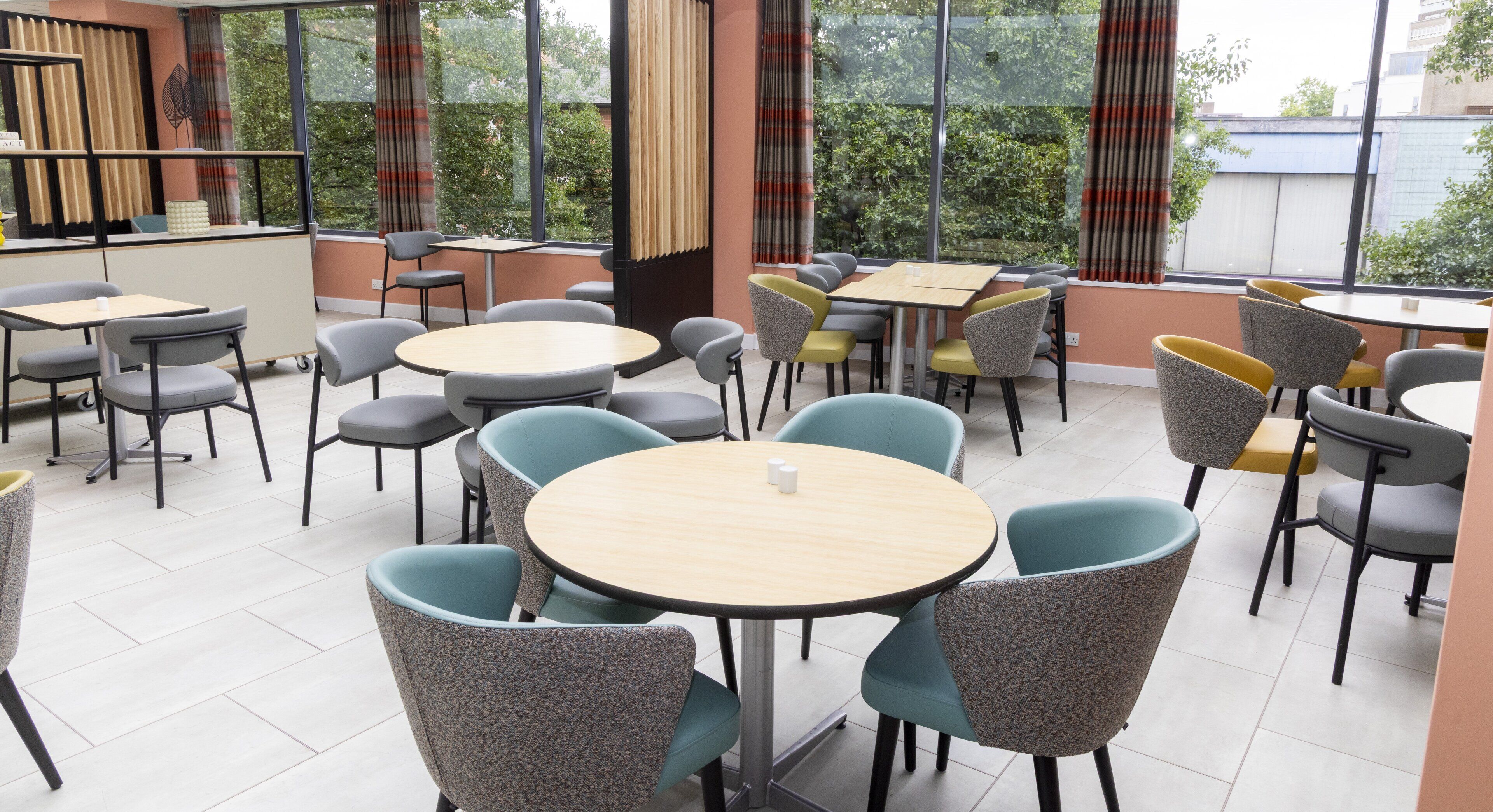 A modern dining area with round tables and chairs, large windows with curtains, and a view of greenery outside.
