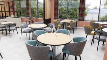 A modern dining area with round tables and chairs, large windows with curtains, and a view of greenery outside.