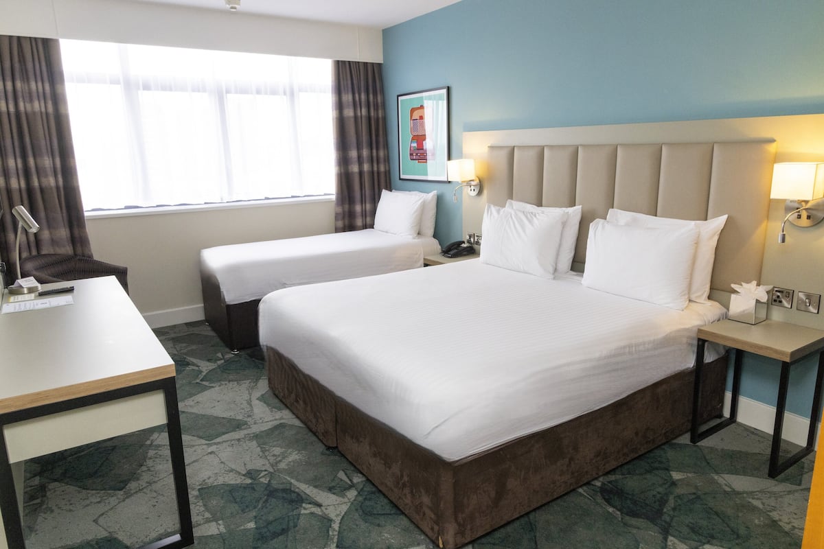 Une chambre d’hôtel avec deux lits simples, un bureau, une chaise, une table de chevet et une fenêtre avec des rideaux.