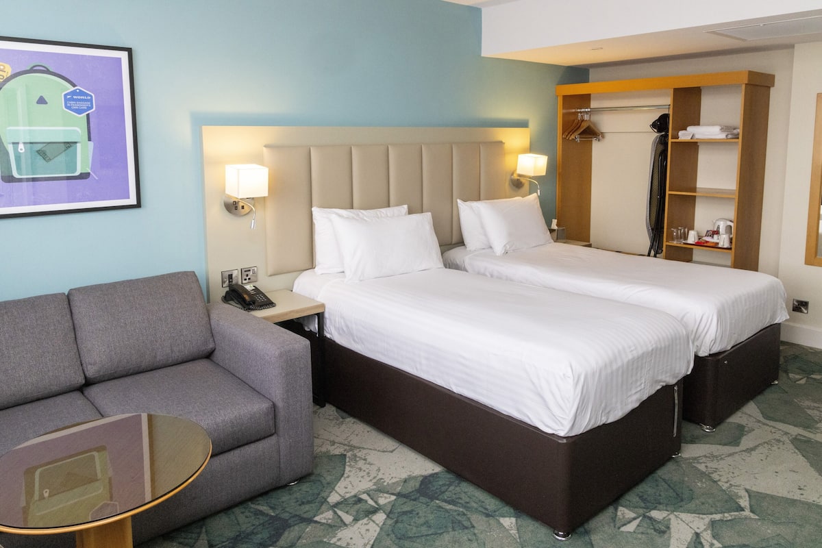 Une chambre d’hôtel avec deux lits, un canapé, une petite table, un porte-manteau et une armoire.