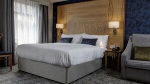 A hotel room with a large bed, two bedside lamps, a wooden headboard, a grey sofa, and a patterned carpet.