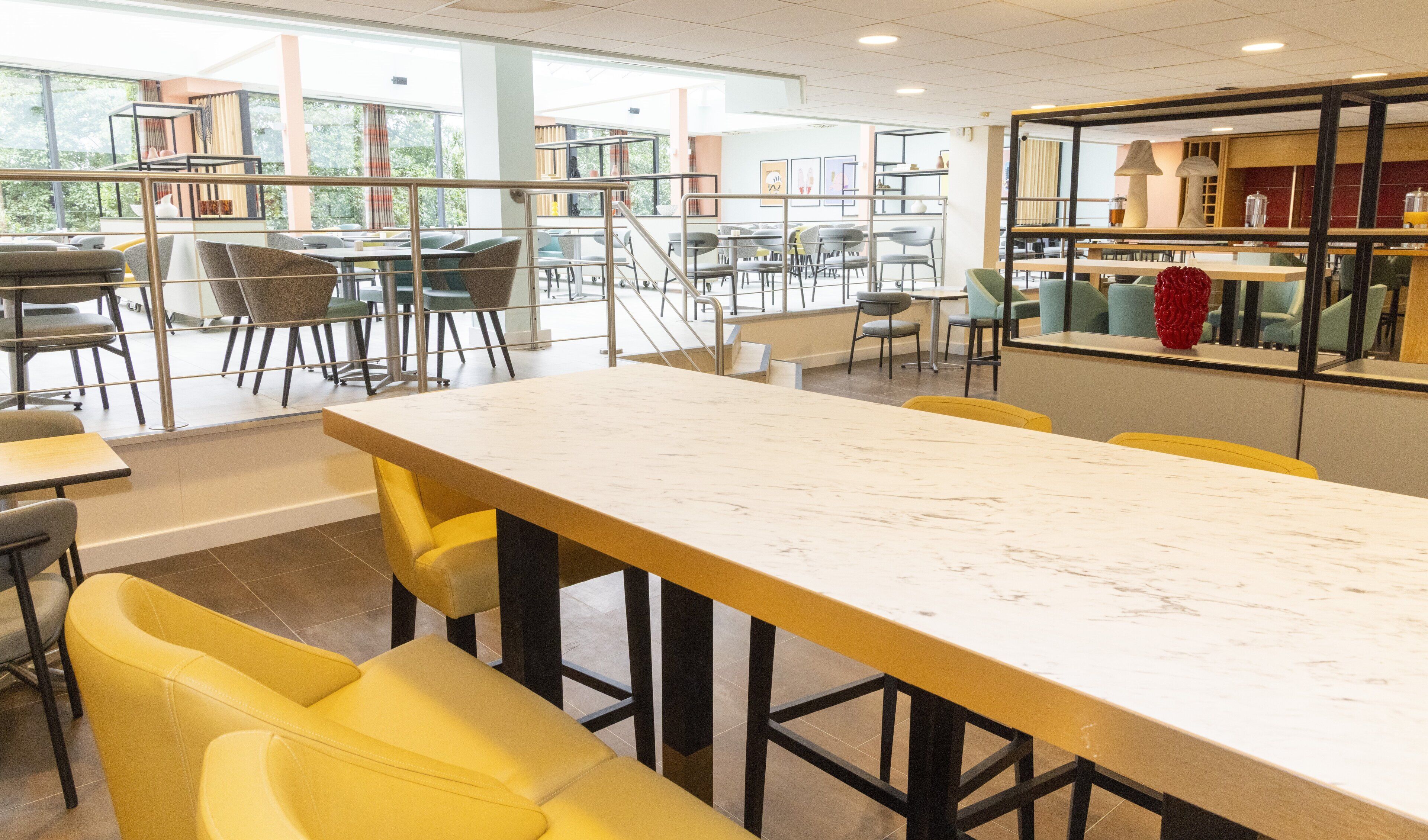 A spacious dining area with high tables and chairs, a staircase leading to an upper level, and large windows offering views of greenery outside.