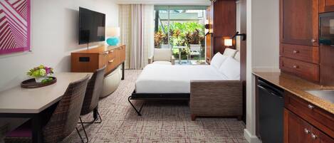 A compact hotel room with a bed, a desk with a chair, a television, a small dining table with chairs, a kitchenette, and a view of a garden through a window.