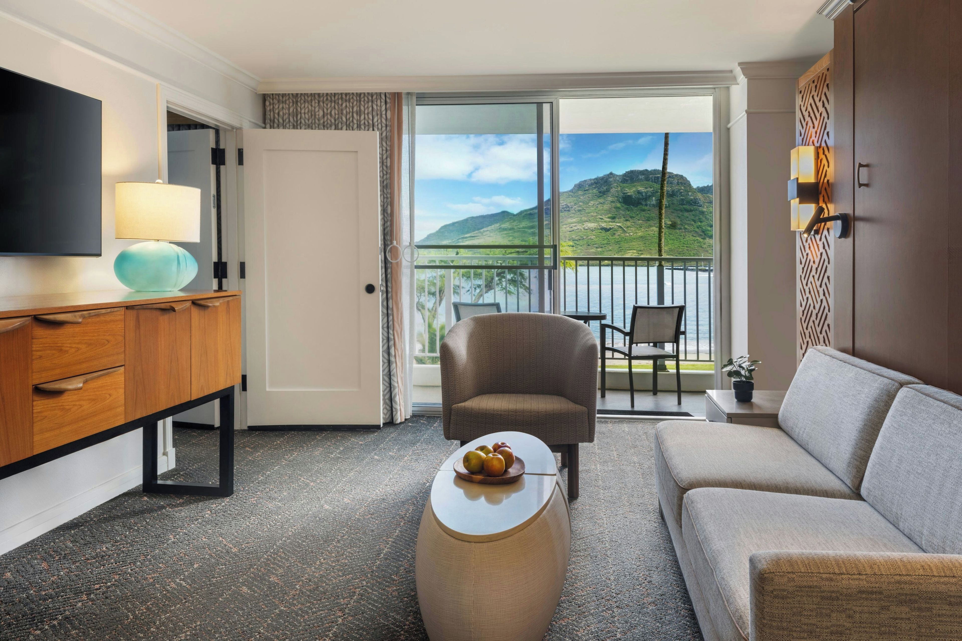 Une chambre d’hôtel équipée d’un canapé, d’un fauteuil, d’une télévision, d’une lampe et offrant une vue sur un balcon donnant sur un paysage pittoresque.