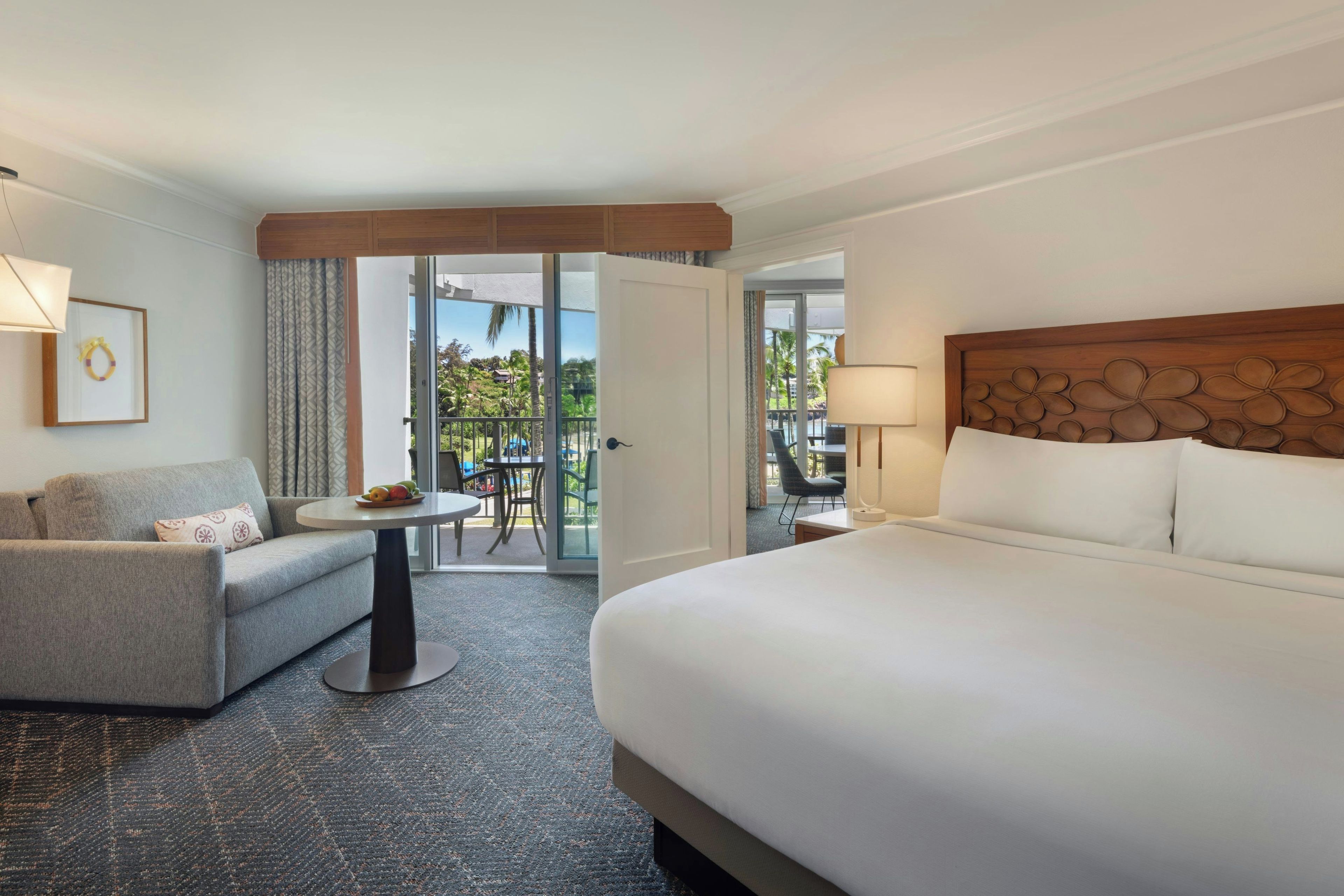 A hotel room with a large bed, a sofa, a small table, and a balcony with a view of palm trees.