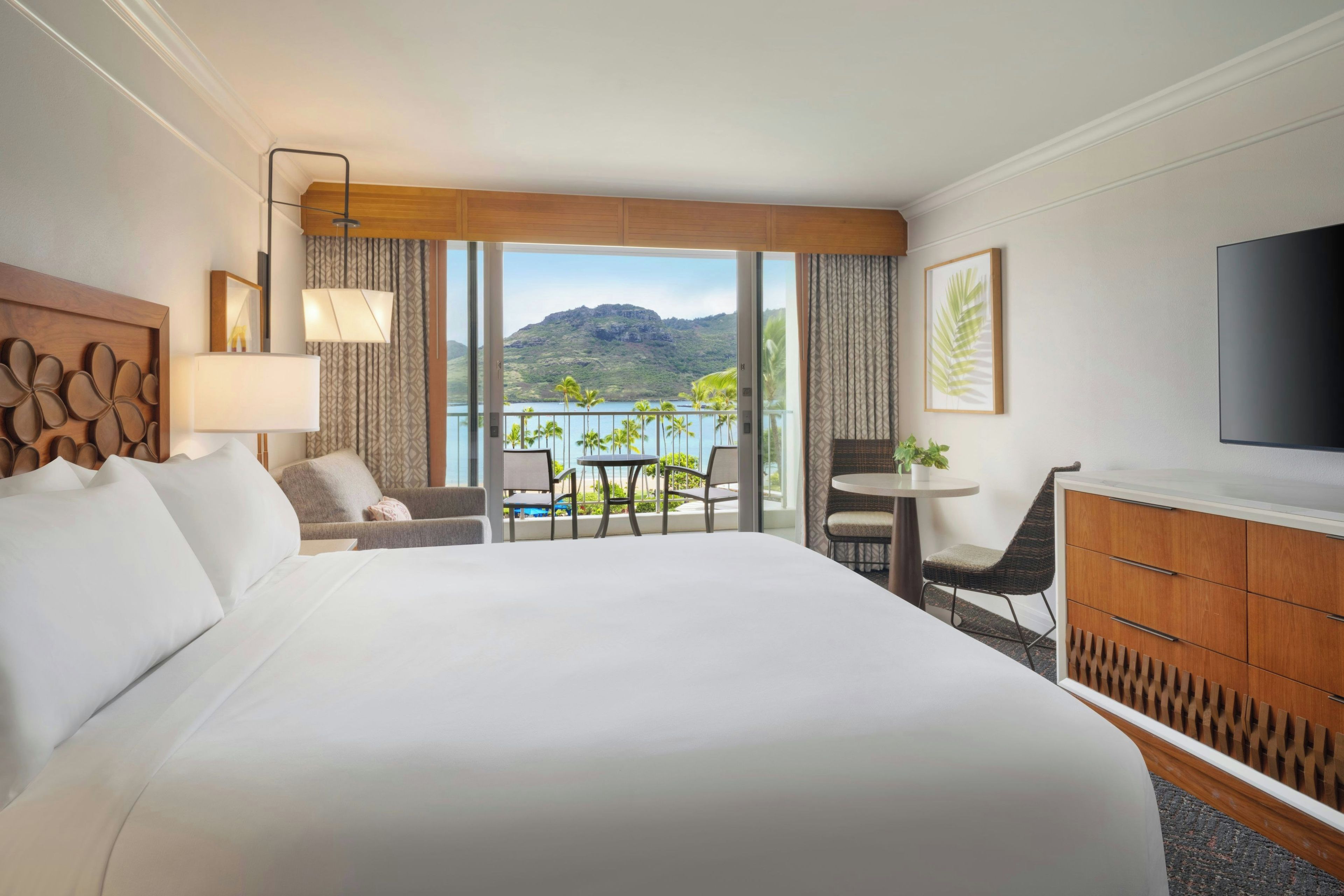 Habitación de hotel con una cama grande, un escritorio con silla, un televisor y un balcón con vista a una montaña y al agua.