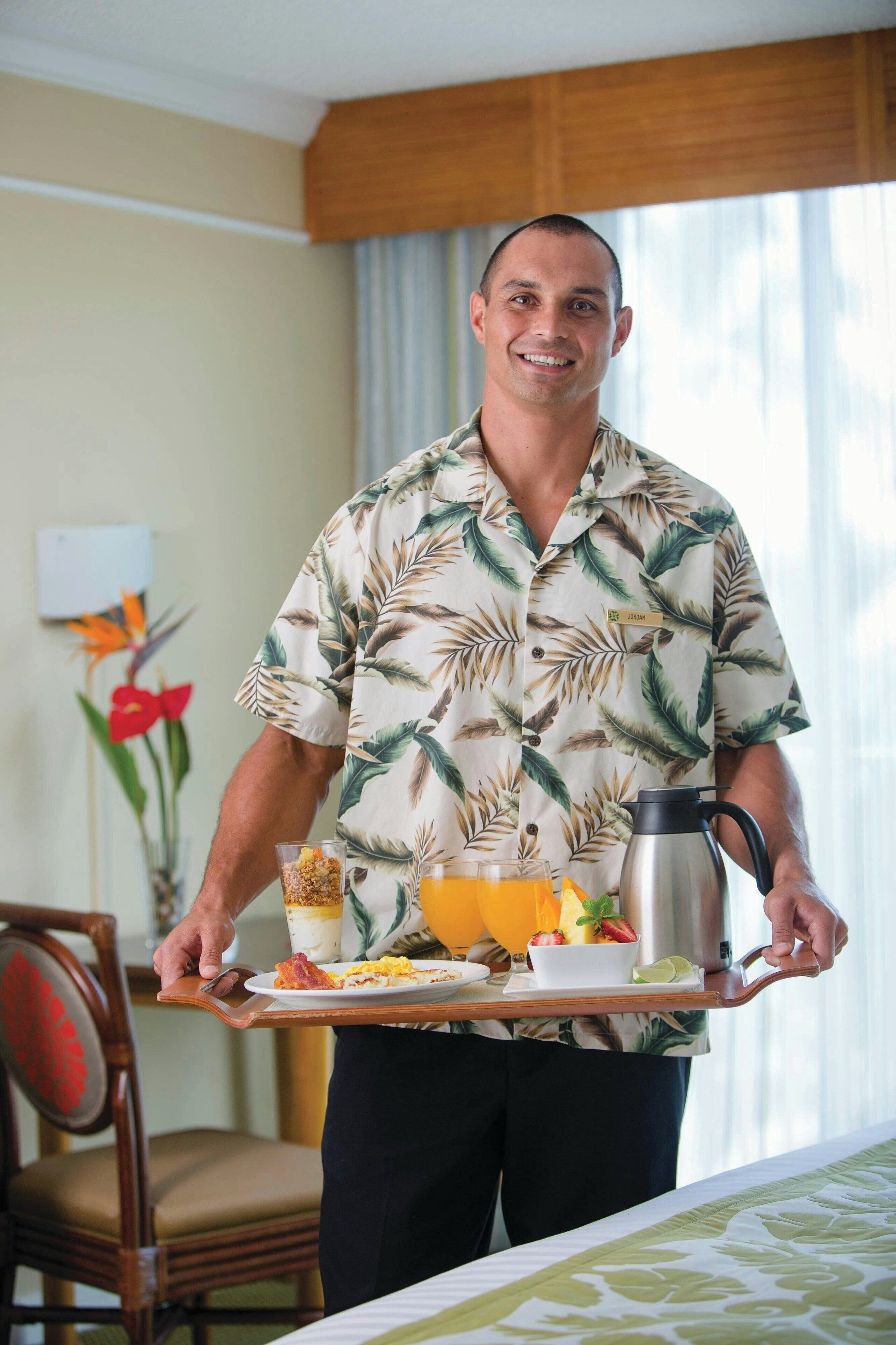 Una persona con camisa de estampado tropical llevando una bandeja con alimentos de desayuno, incluyendo cereal, jugo de naranja y una cafetera.