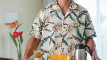 Una persona con camisa de estampado tropical llevando una bandeja con alimentos de desayuno, incluyendo cereal, jugo de naranja y una cafetera.