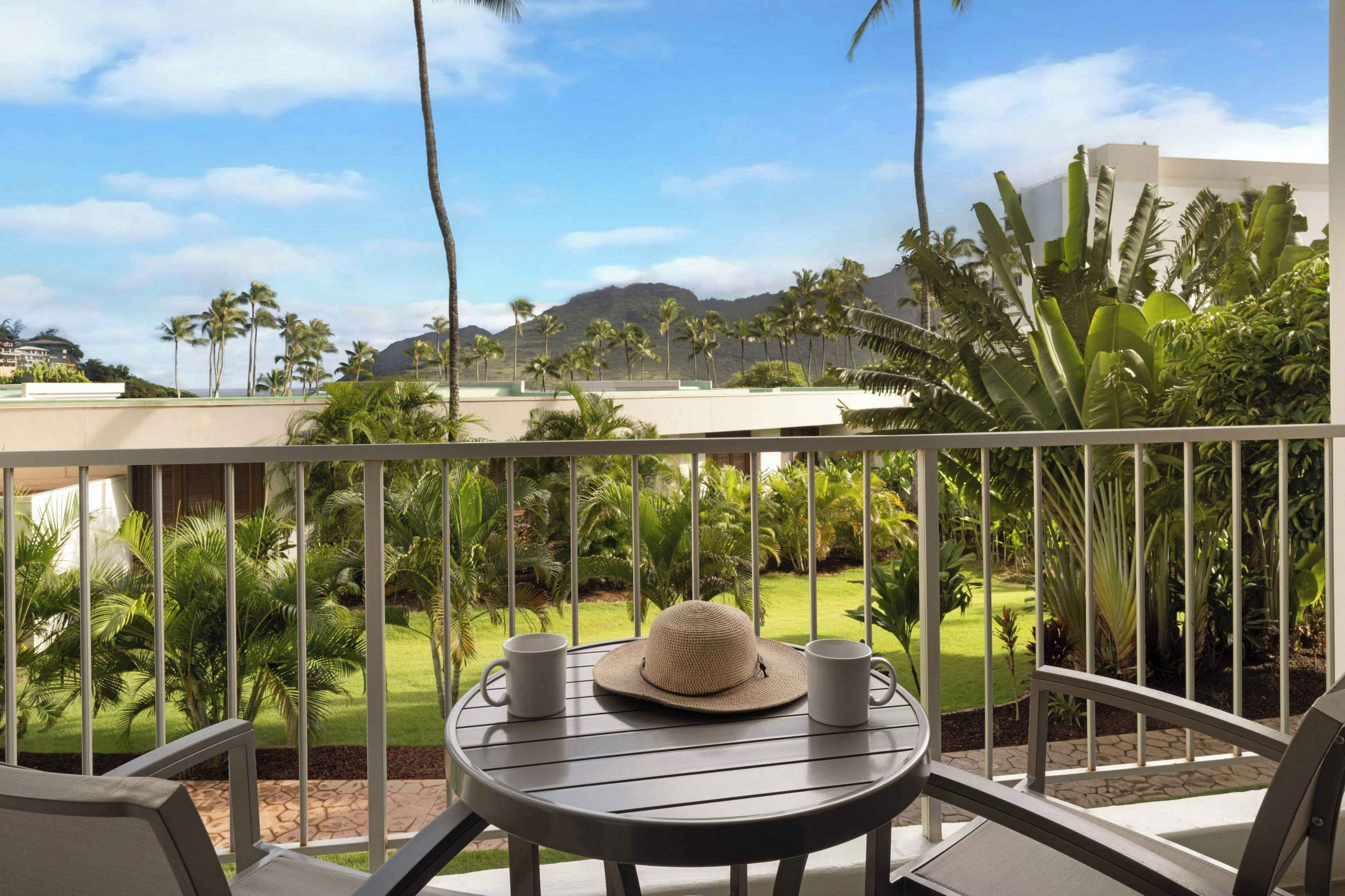 Un balcón con mesa, sillas y vistas a un paisaje tropical con palmeras y montañas.