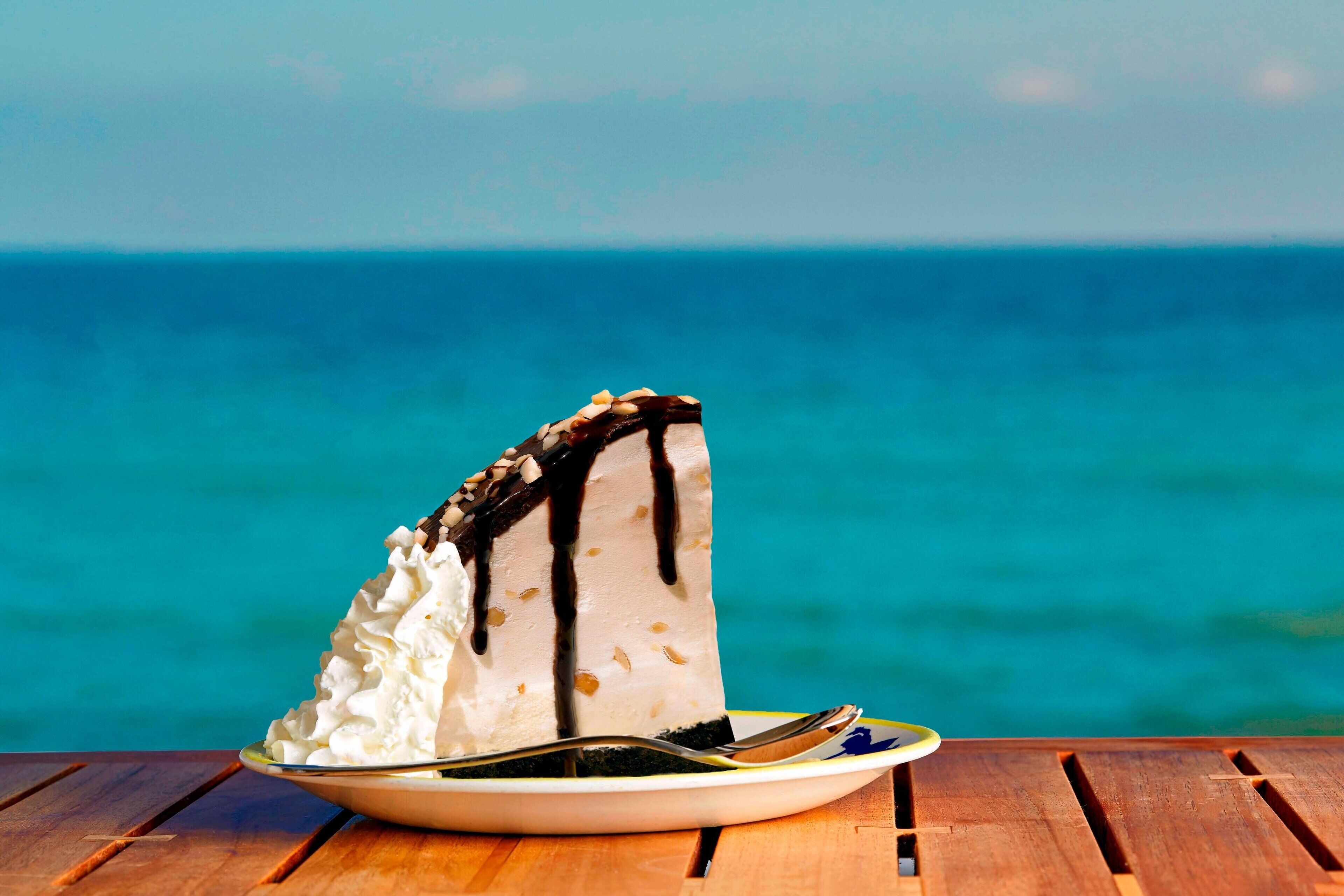 A slice of cake with whipped cream and chocolate drizzle on a plate, placed on a wooden table with a view of the ocean in the background.