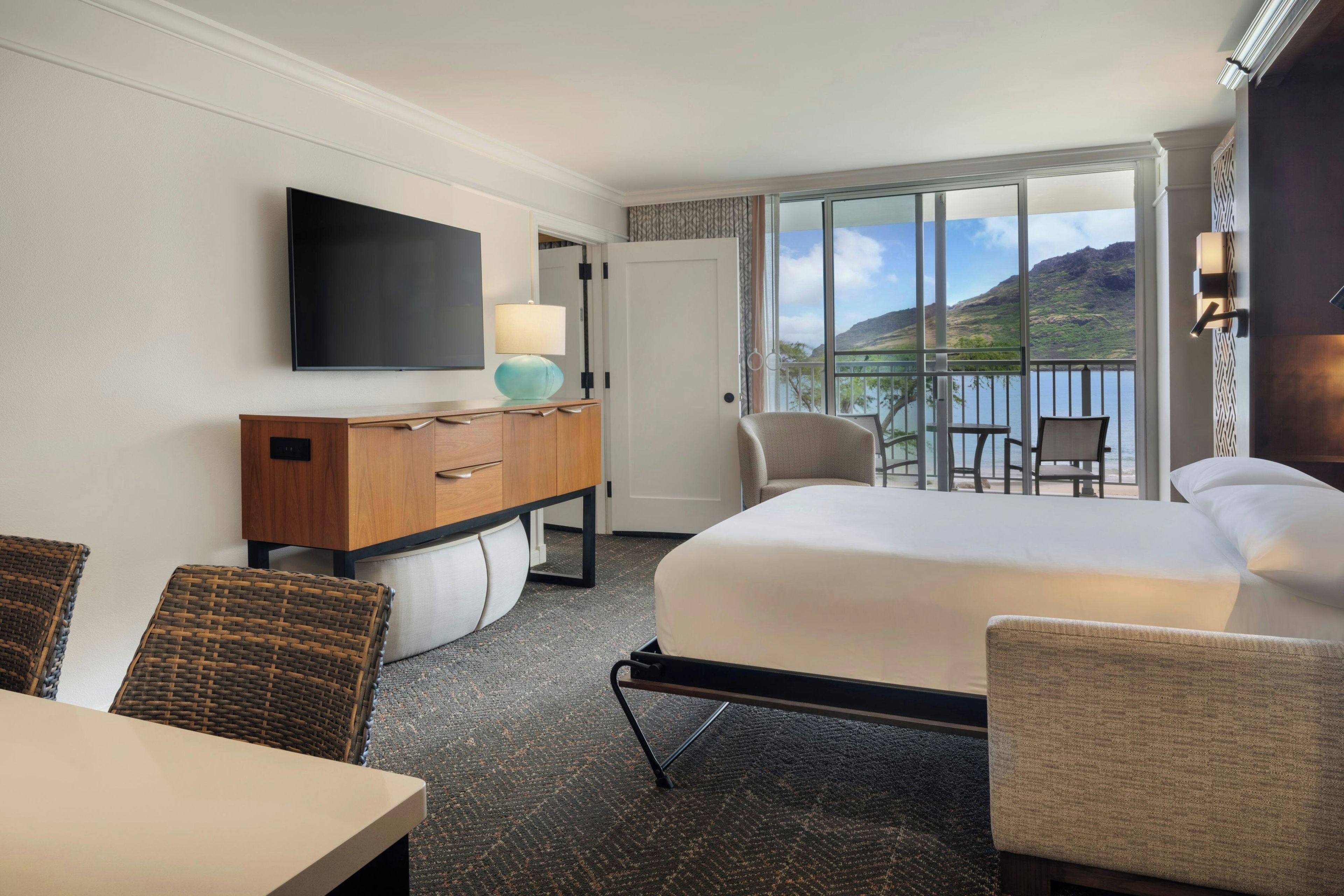 A hotel room with a bed, a desk, a TV, a lamp, a chair, and a balcony with a view of the mountains.