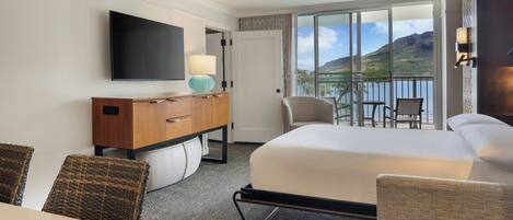 A hotel room with a bed, a desk, a TV, a lamp, a chair, and a balcony with a view of the mountains.